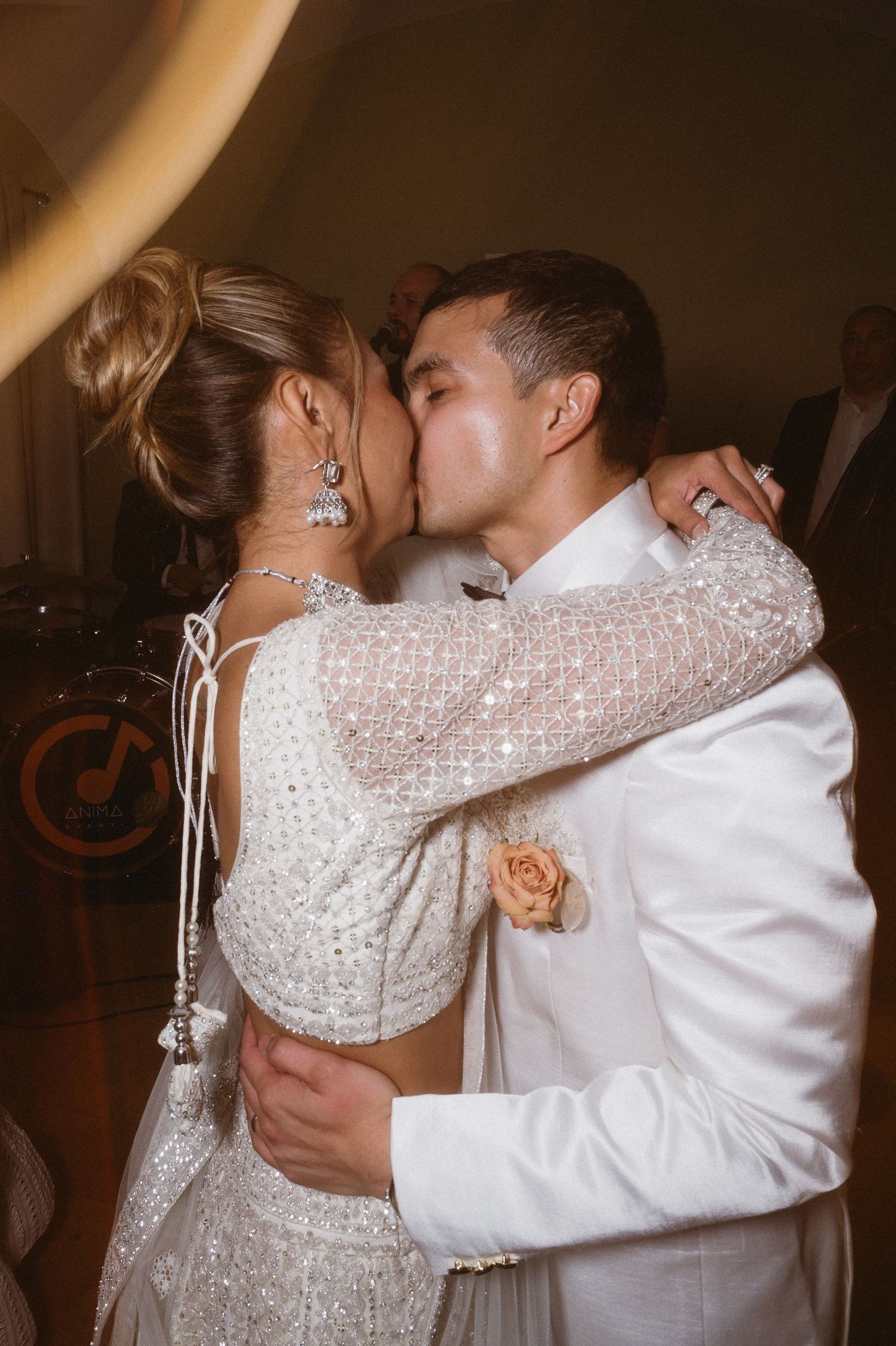 A couple embraces and kisses at a wedding reception, with the bride wearing a lace dress with a peach-colored boutonniere.