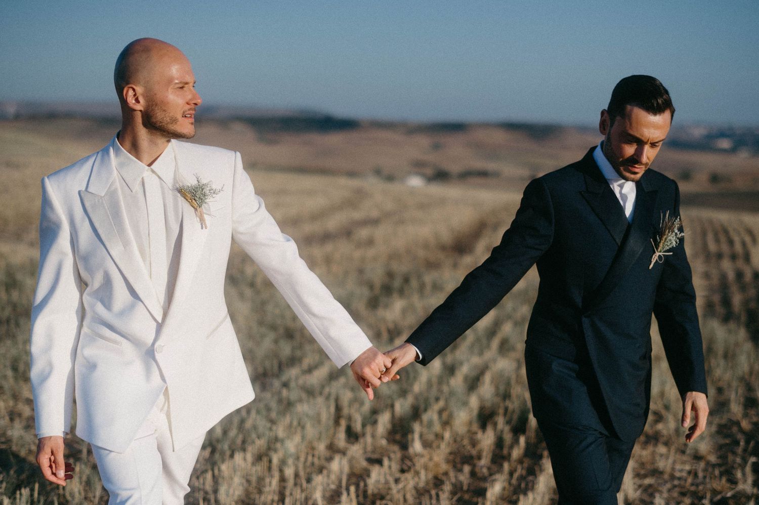 Two people in contrasting white and black suits hold hands while walking through a golden dry grass field at sunset.