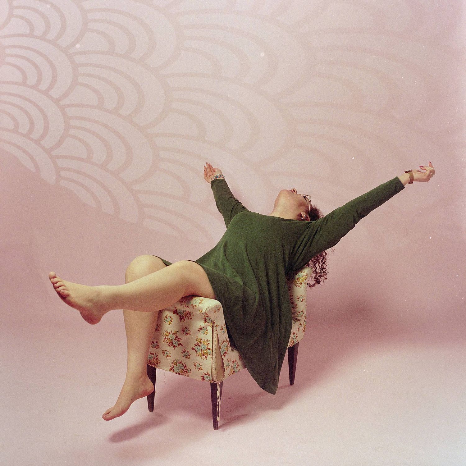 Someone in a green sweater dress lounging carefree on a vintage floral chair against a pink background with scalloped pattern.