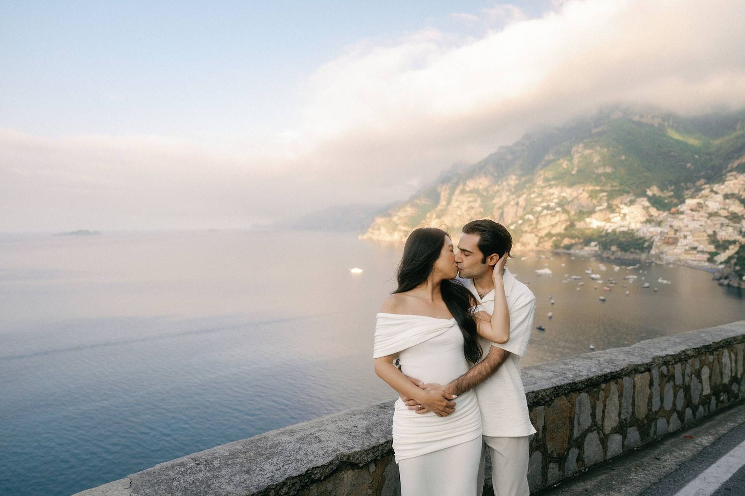 A romantic couple in white outfits embraces on a scenic overlook above the Amalfi Coast in Italy.