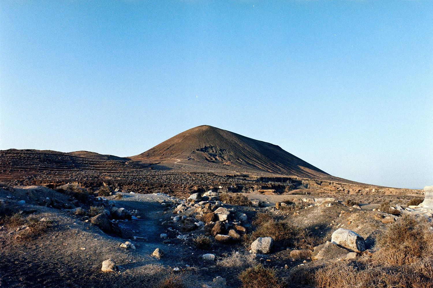 A barren desert landscape with scattered volcanic rocks and a brown mountain peak against a clear blue sky.