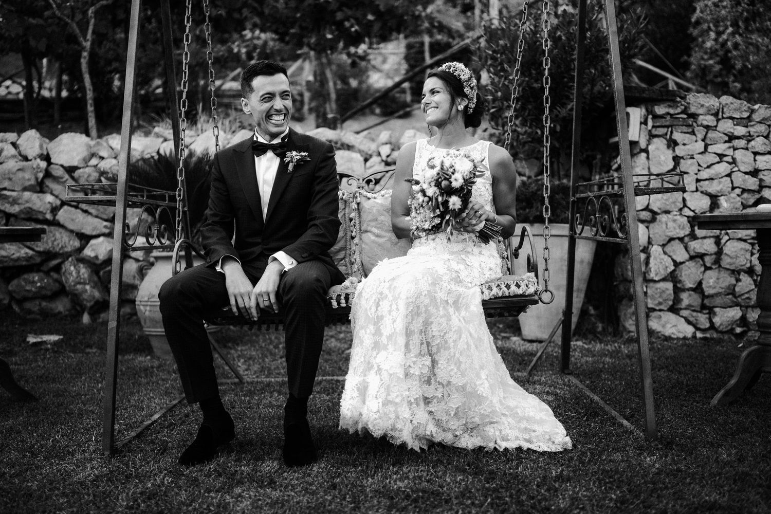 A bride in a lace wedding gown and groom in a tuxedo sit together on a wooden swing against a stone wall.