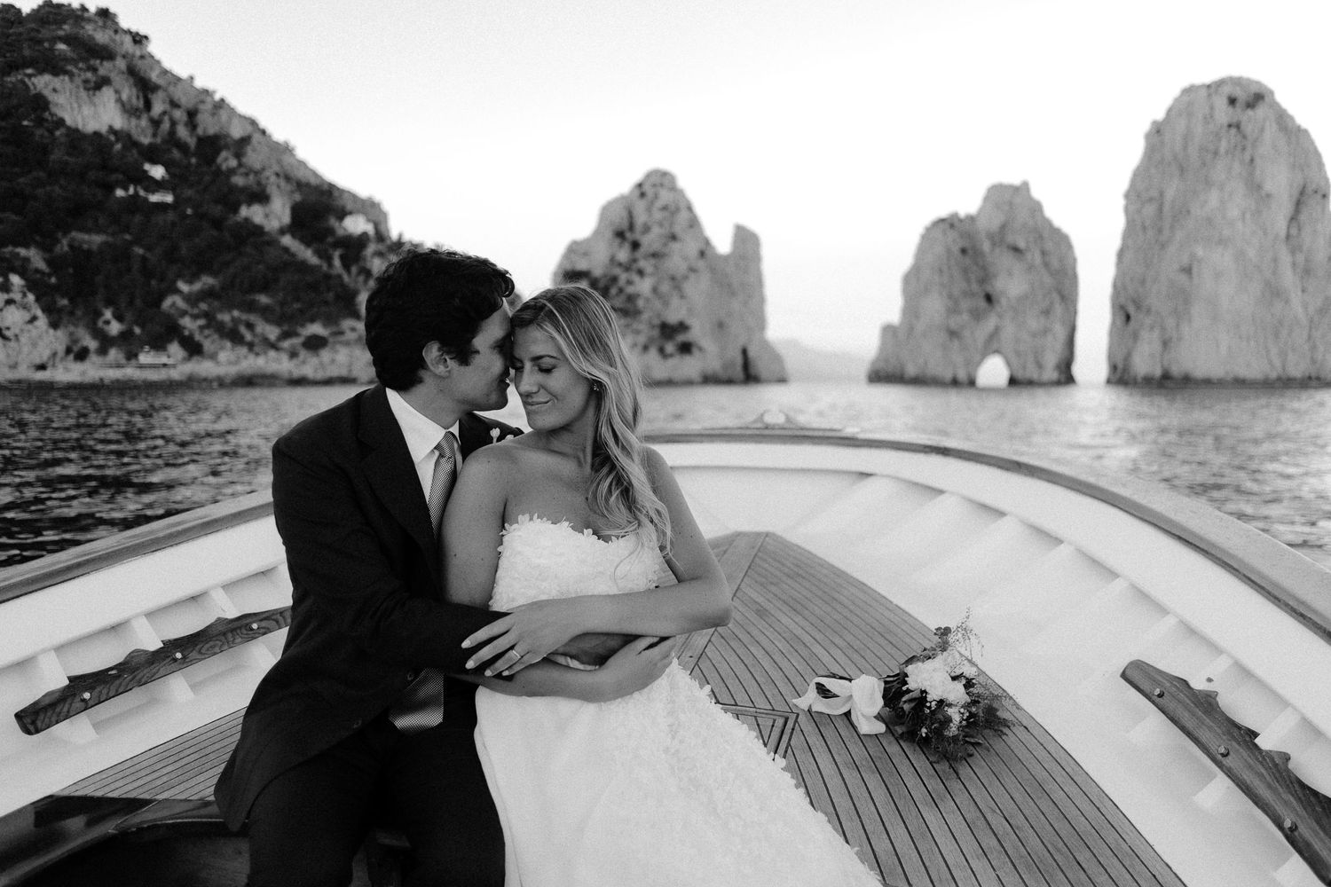 A couple share an intimate moment on a boat deck with dramatic rocky cliffs of Capri in the background.