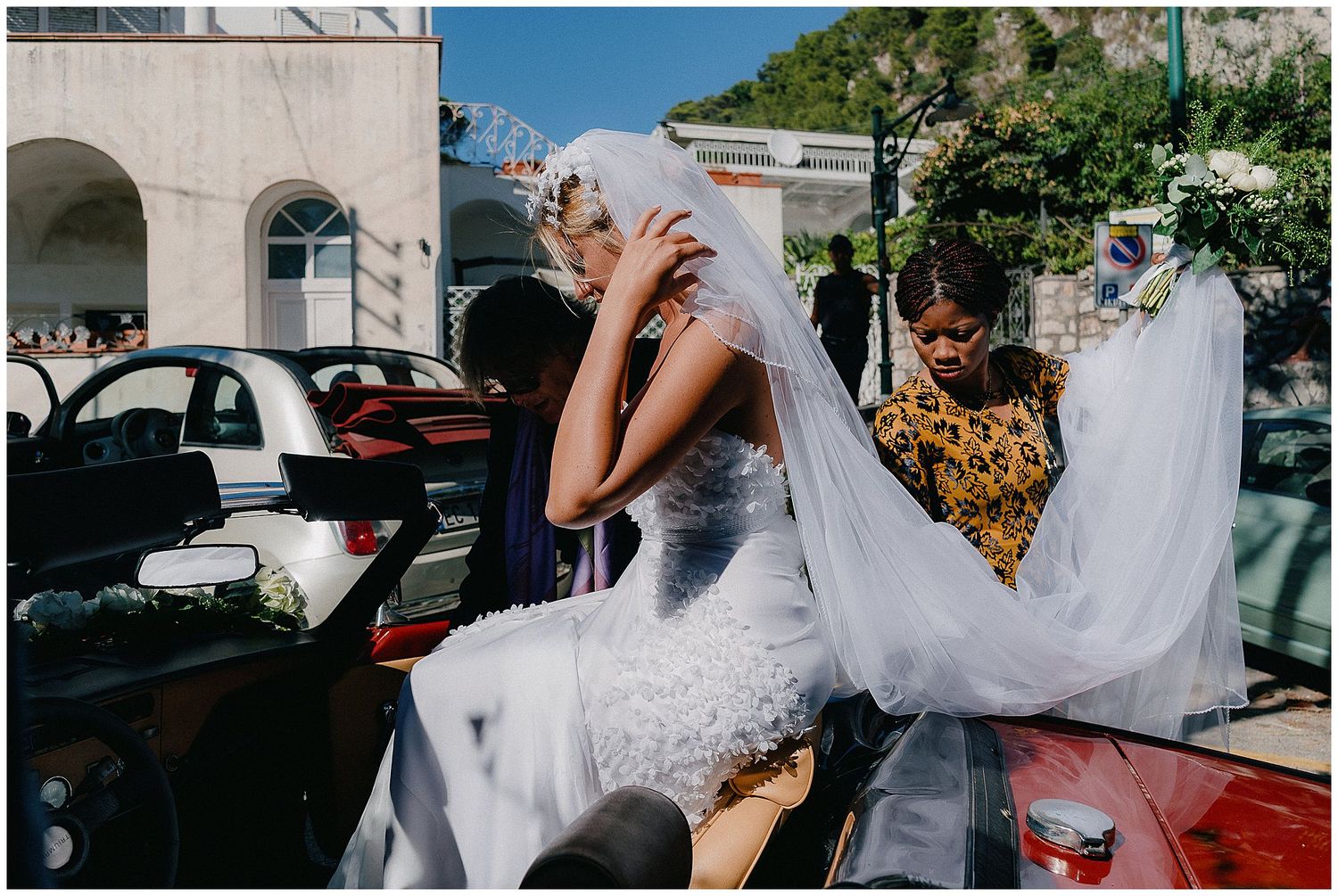 A person in a flowing white wedding dress veil sits in a vintage car on a sunny street with buildings in background.