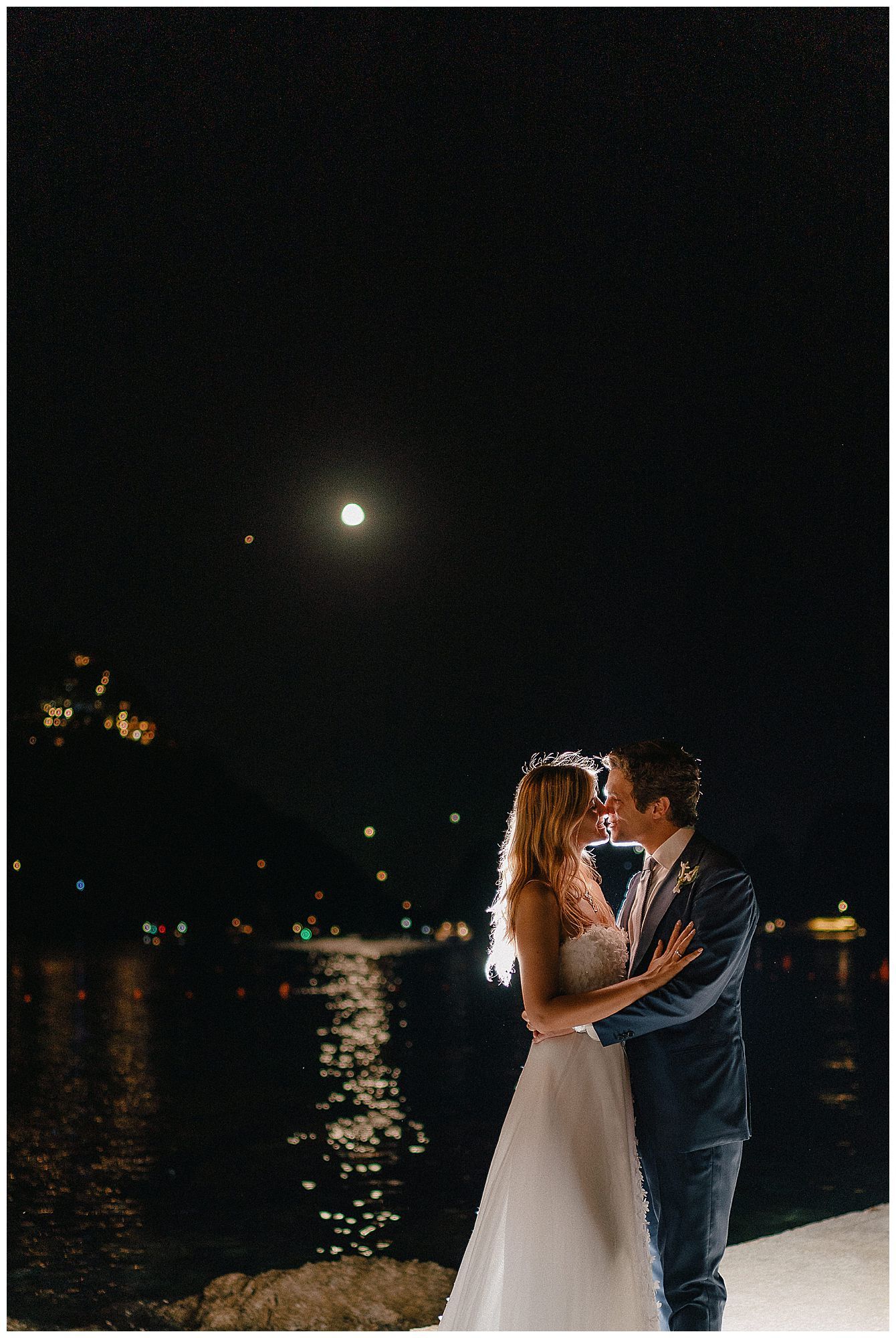 A romantic moonlit couple stands by a waterfront at night with the moon's reflection glimmering on the dark water.