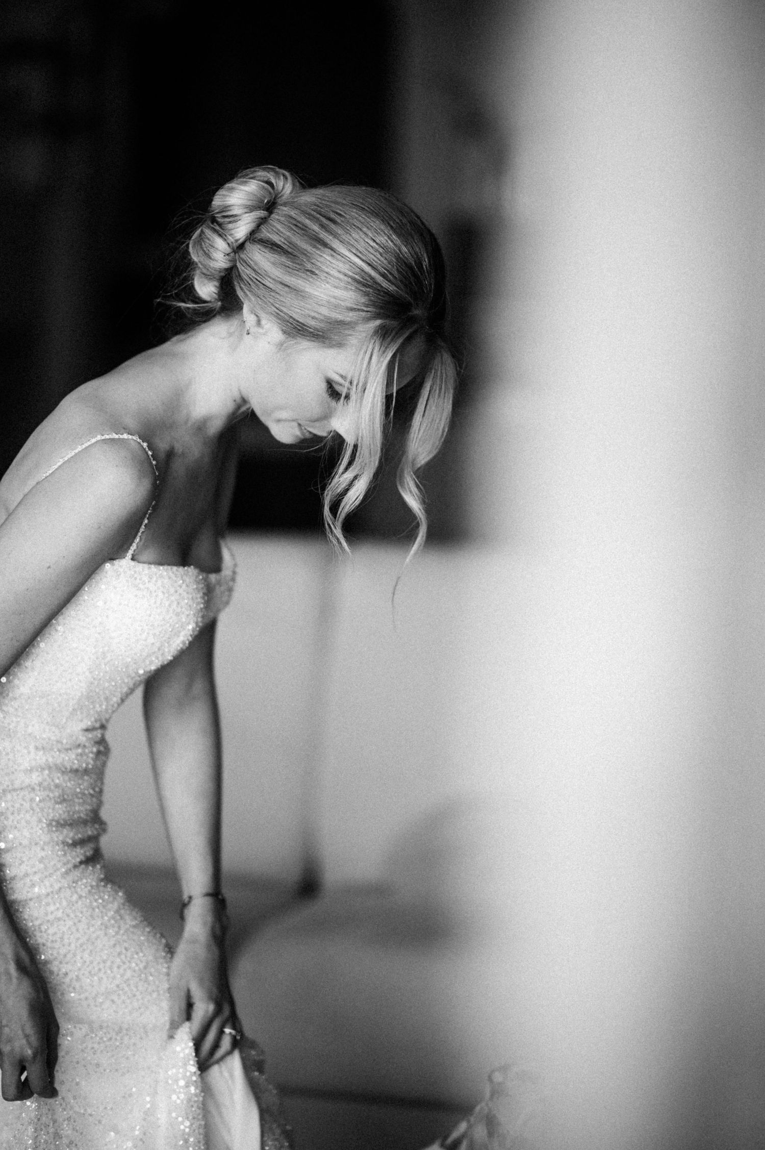 Elegant black and white photograph of a bride in a beaded form-fitting gown adjusting her dress in a quiet moment.