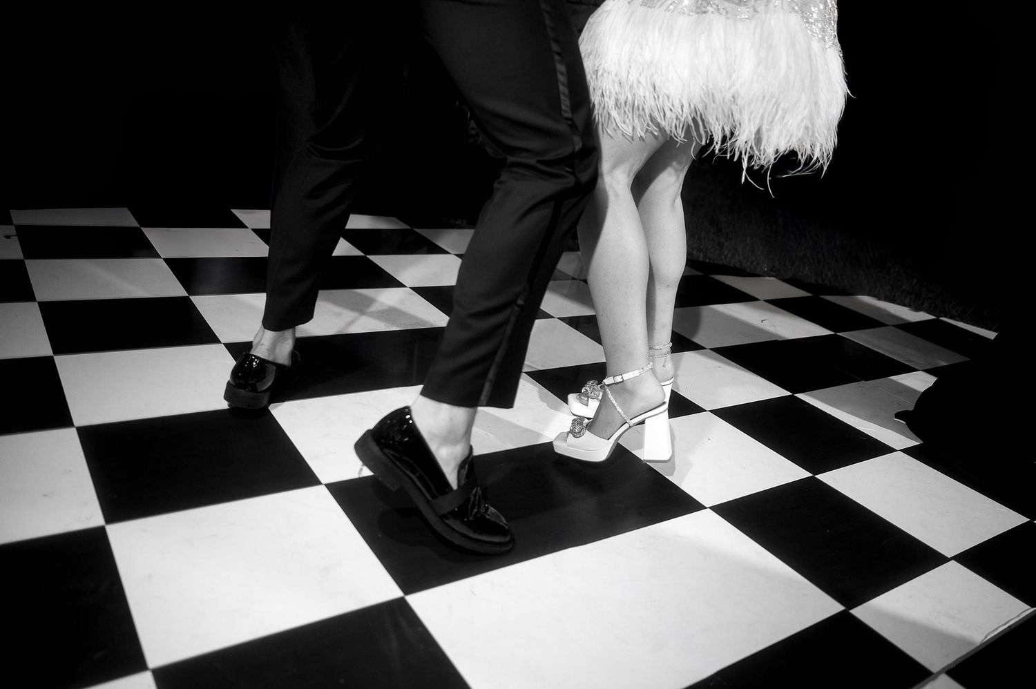 Dancers' feet in black shoes perform on a striking black and white checkered dance floor pattern.