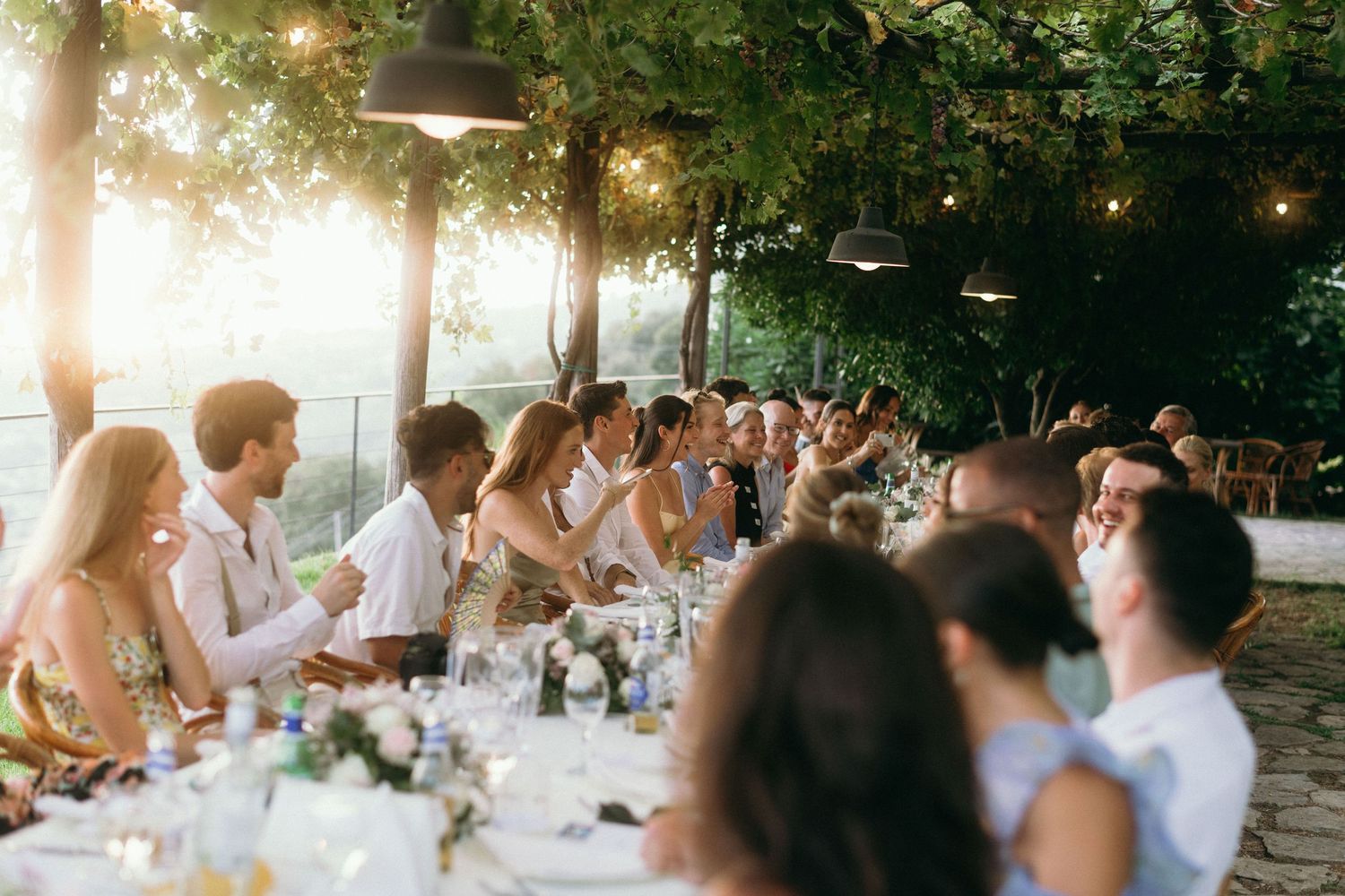 Guests enjoying an elegant outdoor dinner party at long tables under string lights and tree canopy at dusk.
