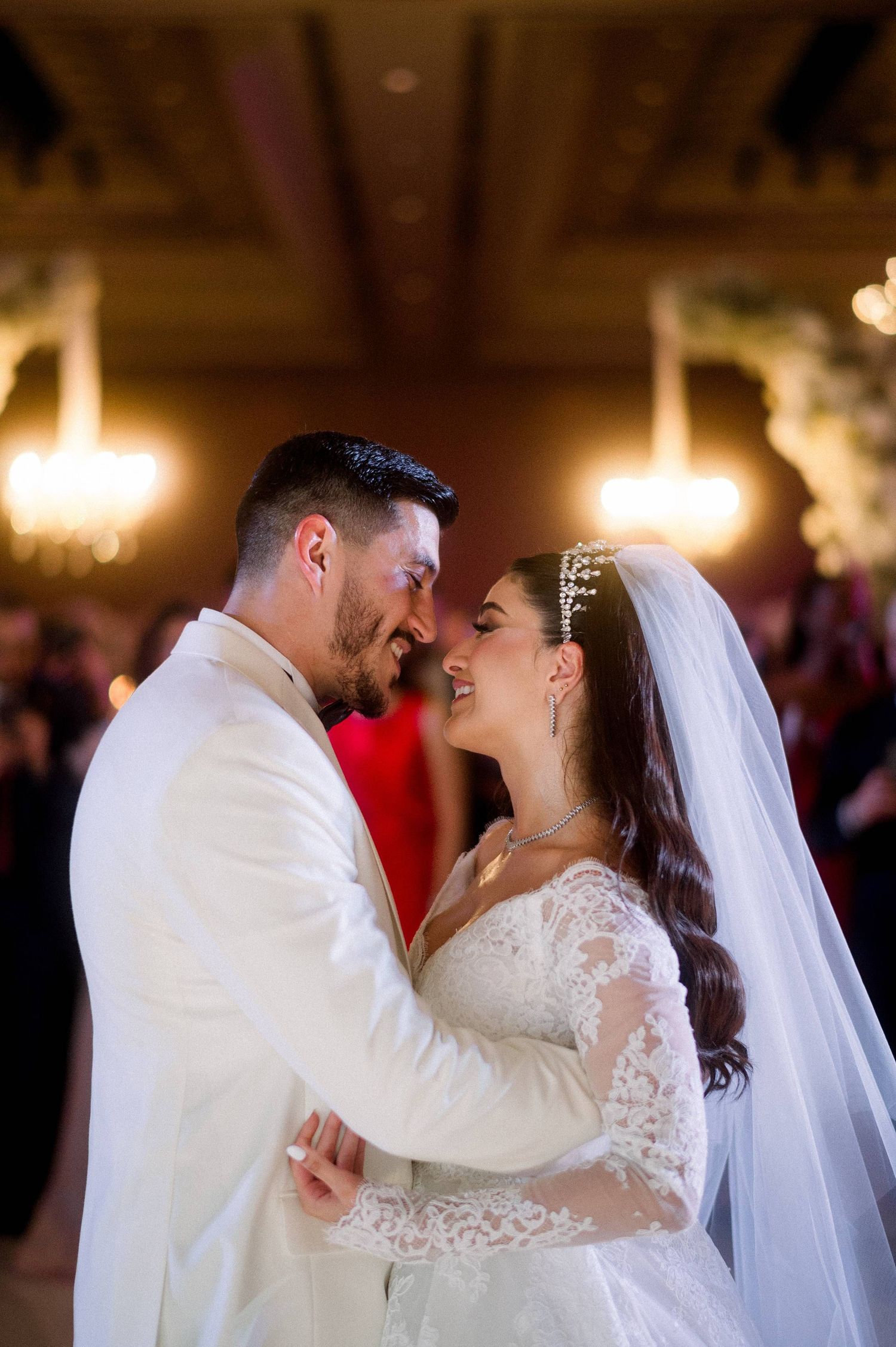 A romantic wedding couple shares an intimate moment on the dance floor under warm chandelier lighting.