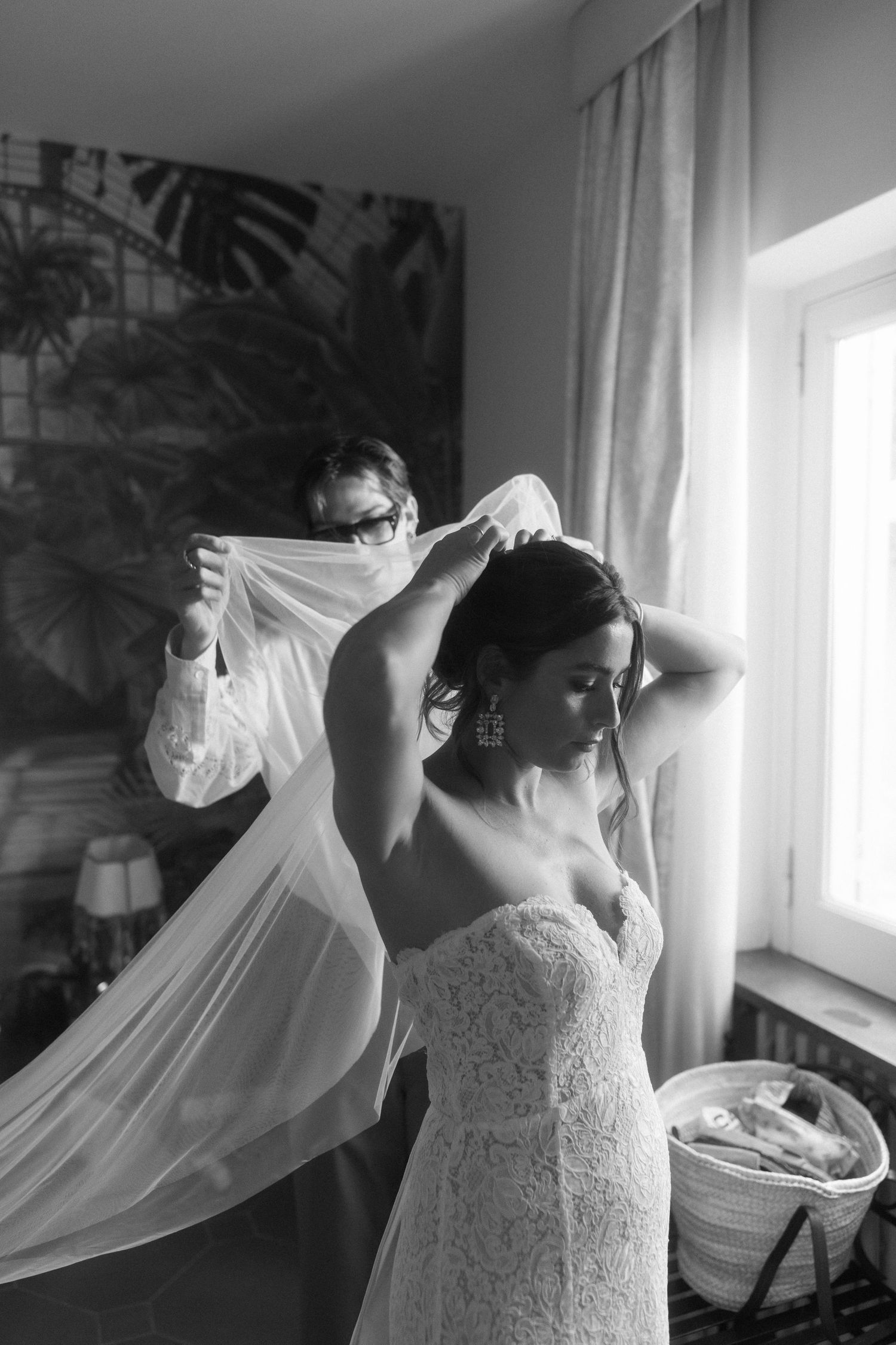 A bride in a white lace wedding dress adjusts her flowing veil near a sunlit window in a black and white photograph.