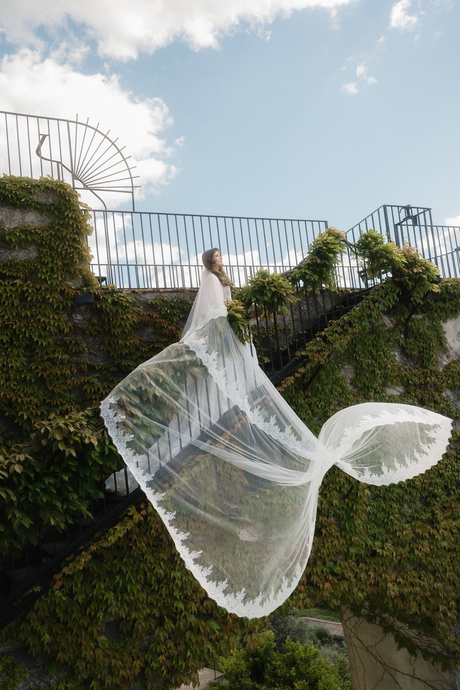 Bride in white wedding dress with flowing veil caught by wind on ivy-covered stone staircase under blue sky.