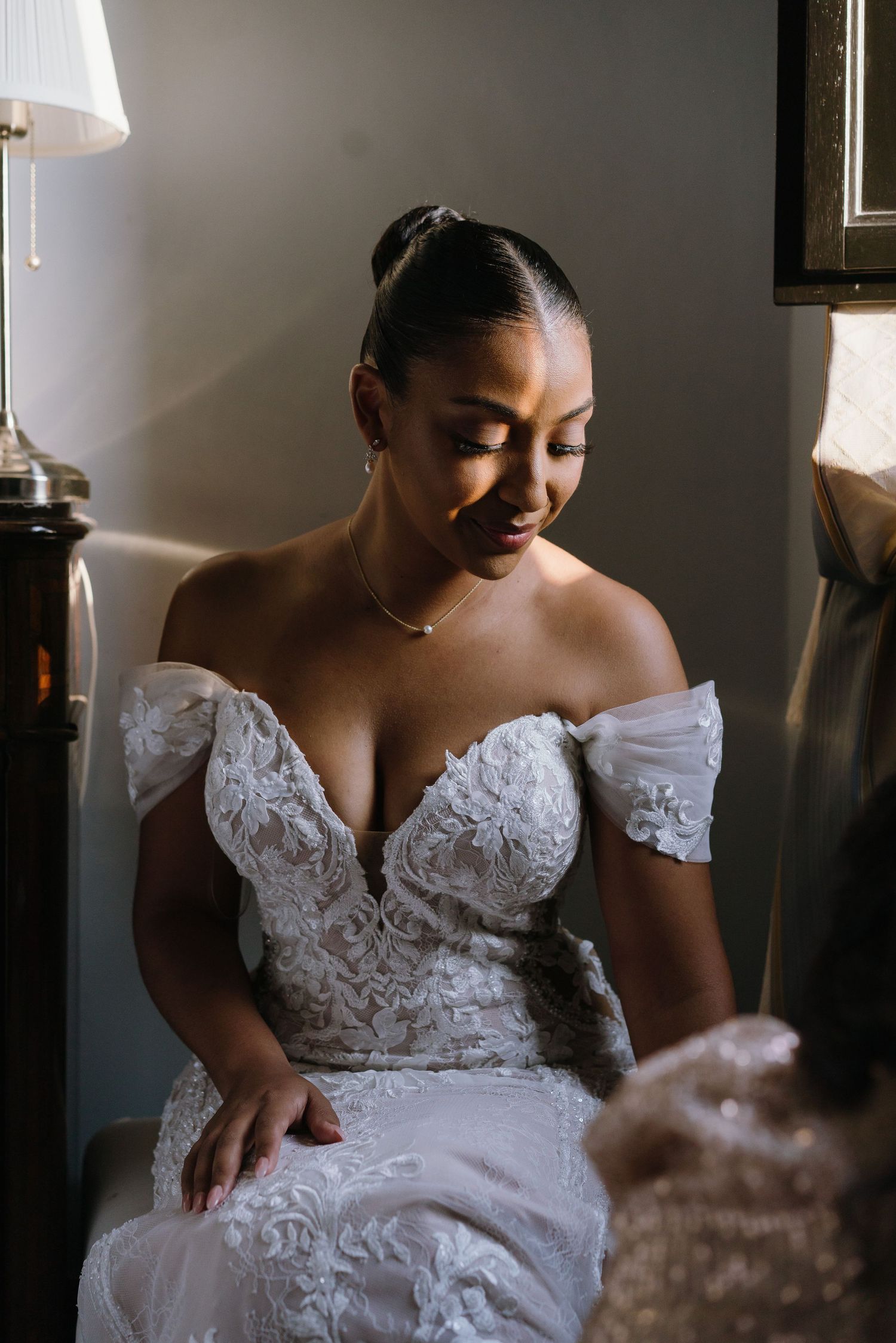 A bride in an off-shoulder lace wedding gown with delicate details sits gracefully in soft lighting.