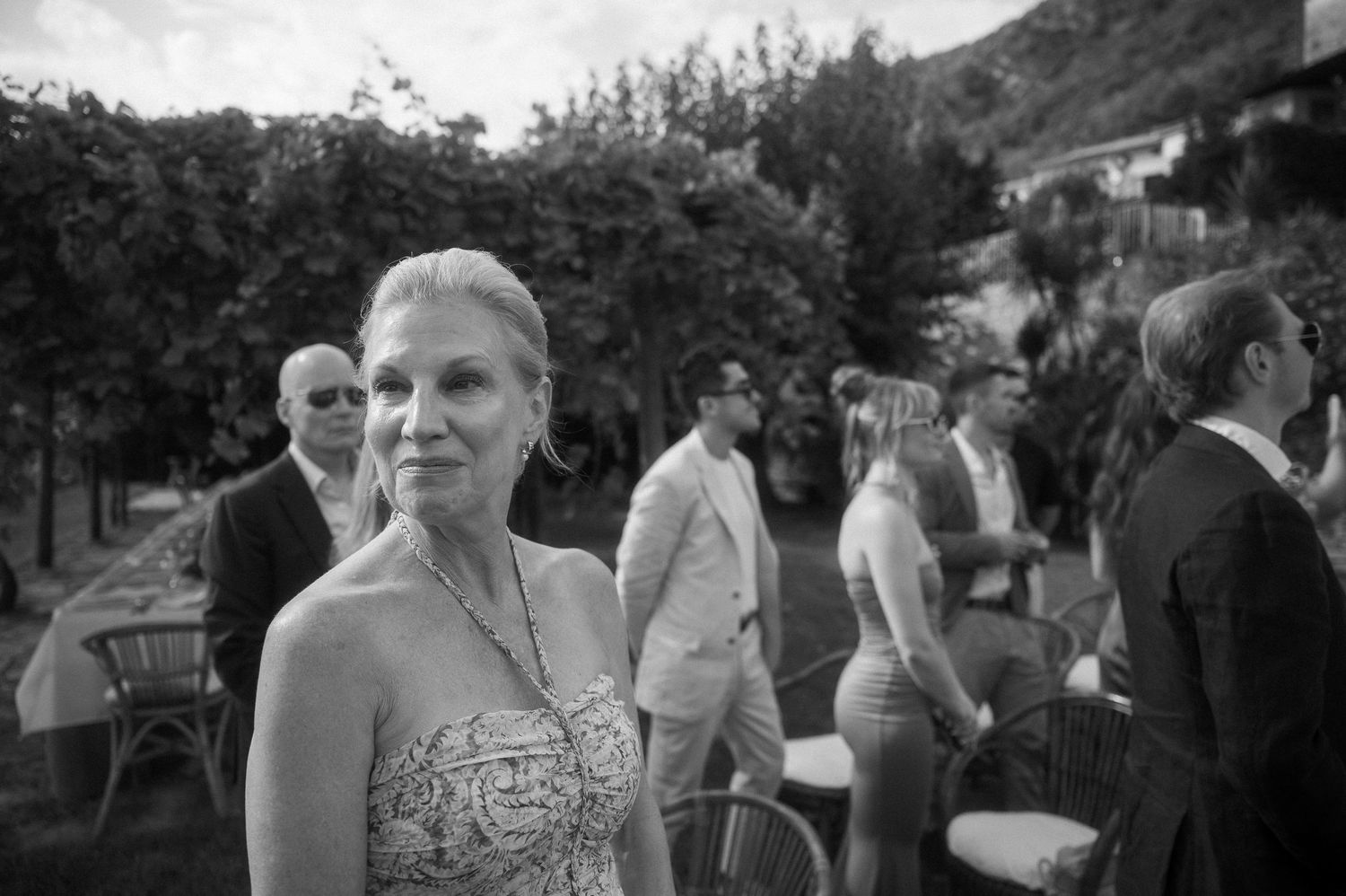 Black and white image of people at an outdoor social gathering with trees and hillside in the background.
