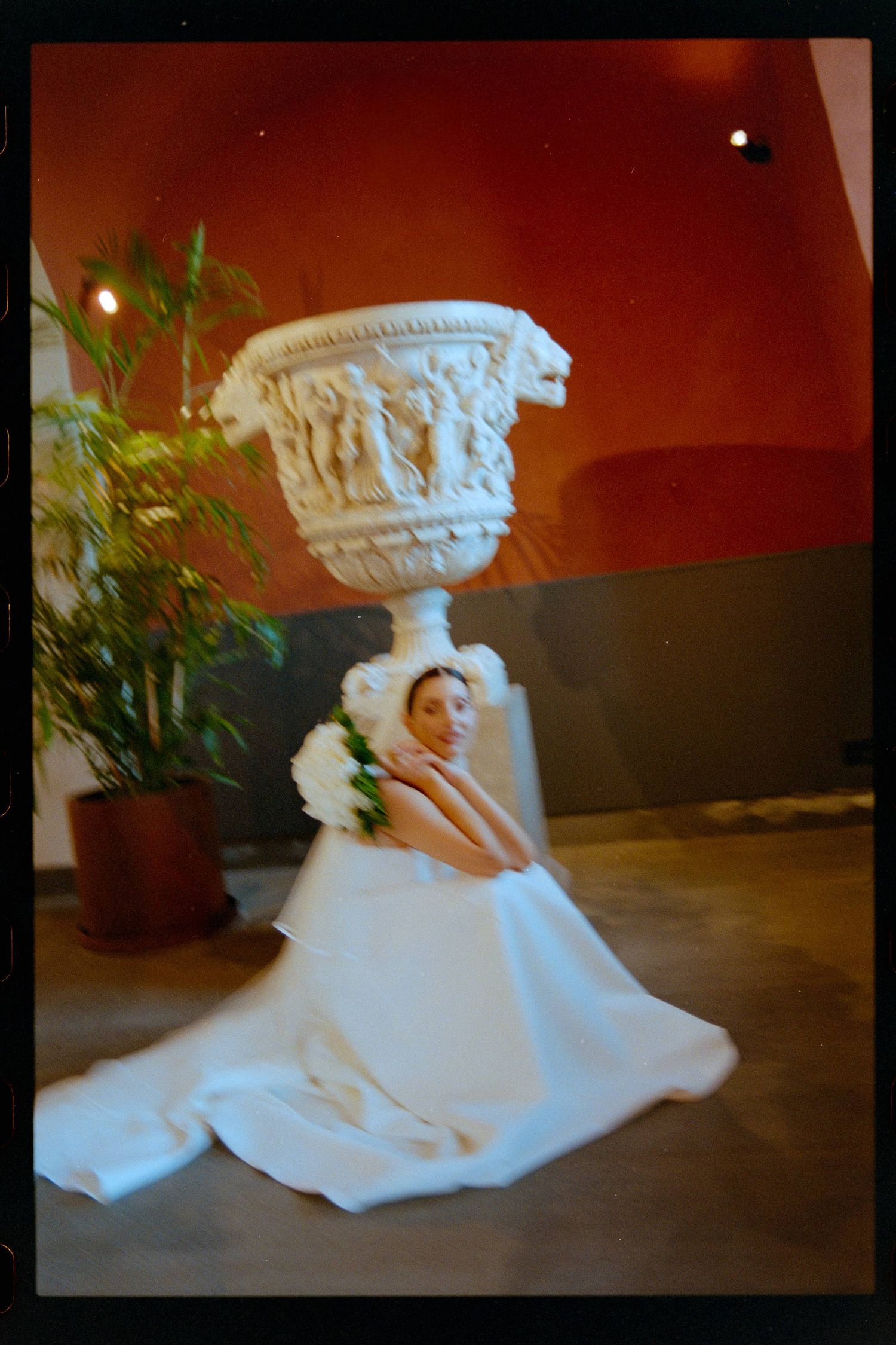 A bride in a white gown poses gracefully beneath an ornate stone urn pedestal, holding a bouquet indoors.