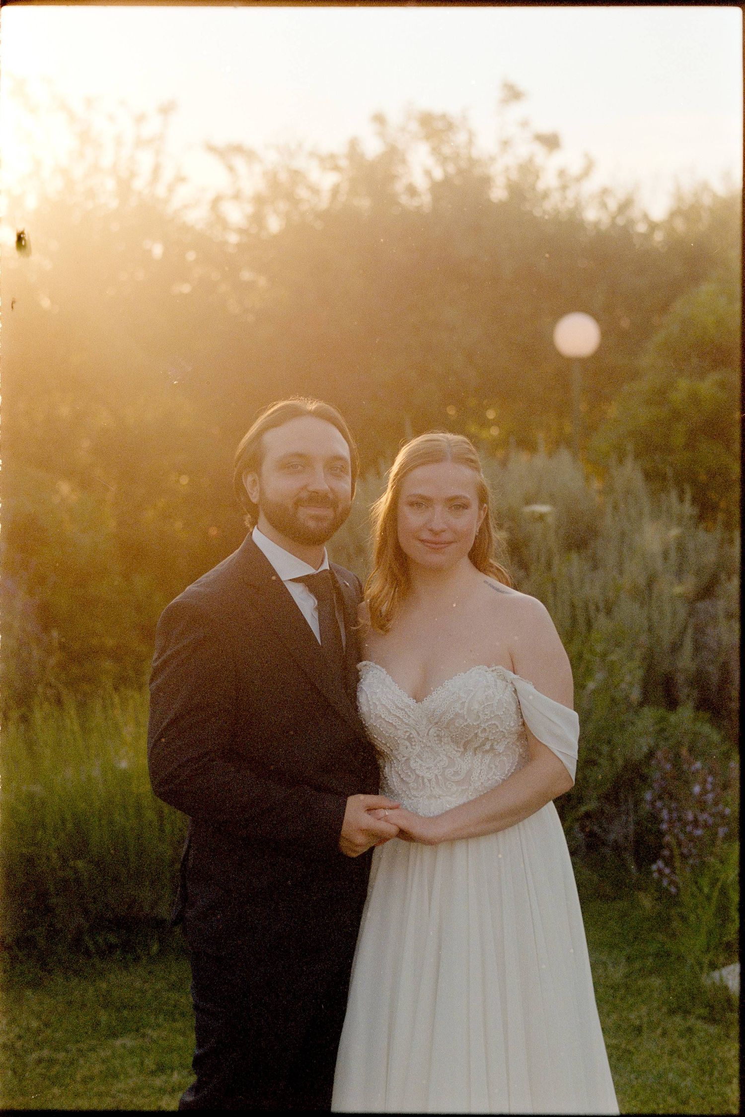 A couple poses together outdoors at sunset in formal wedding attire with golden light and greenery in the background.