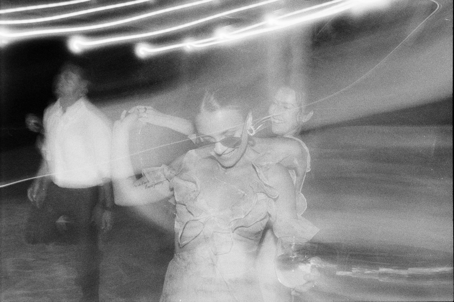 Blurred black and white motion photograph of people dancing at night with streaking light trails.