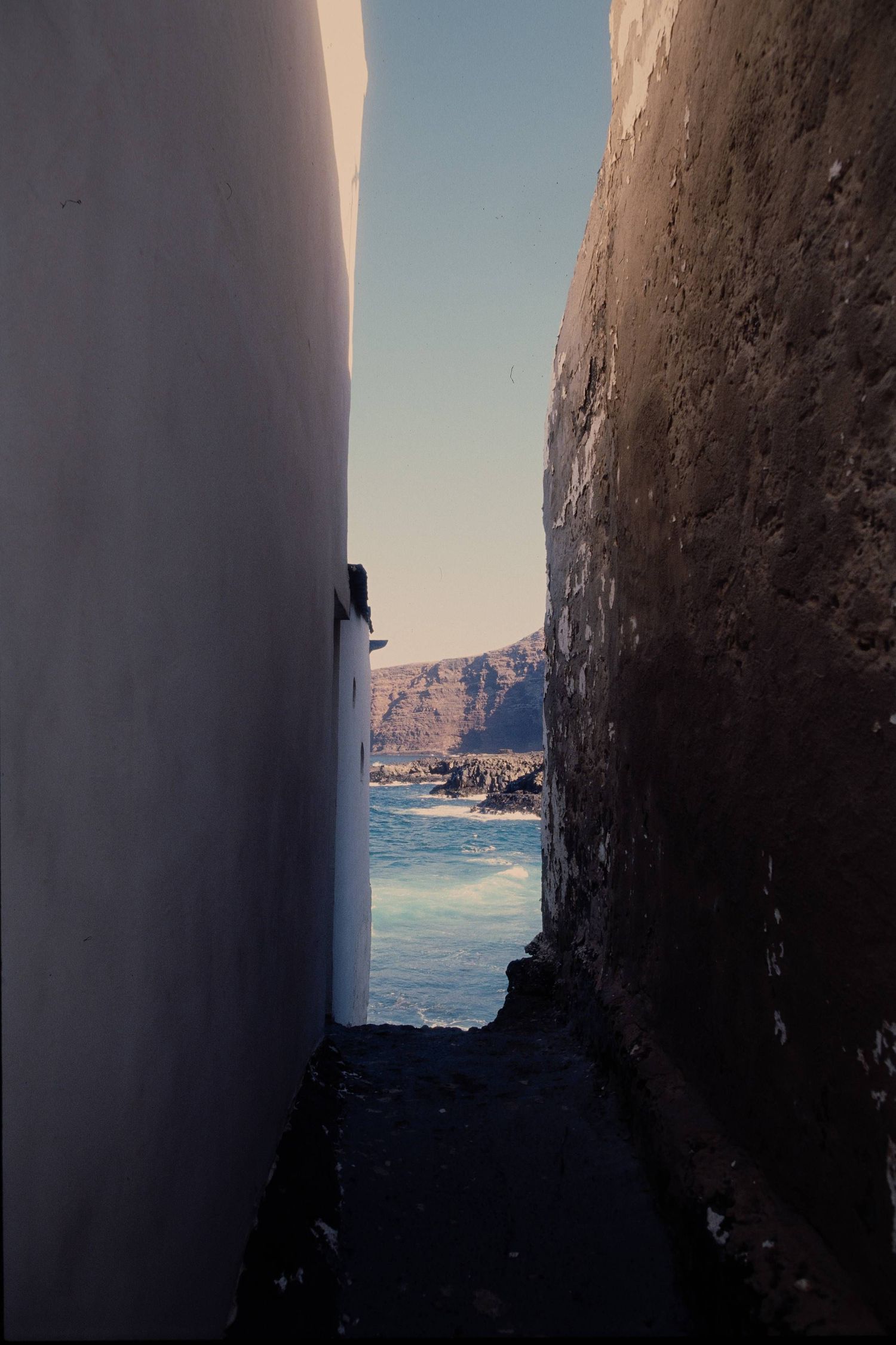 A narrow alleyway between white walls frames a view of coastal cliffs and blue ocean waters.