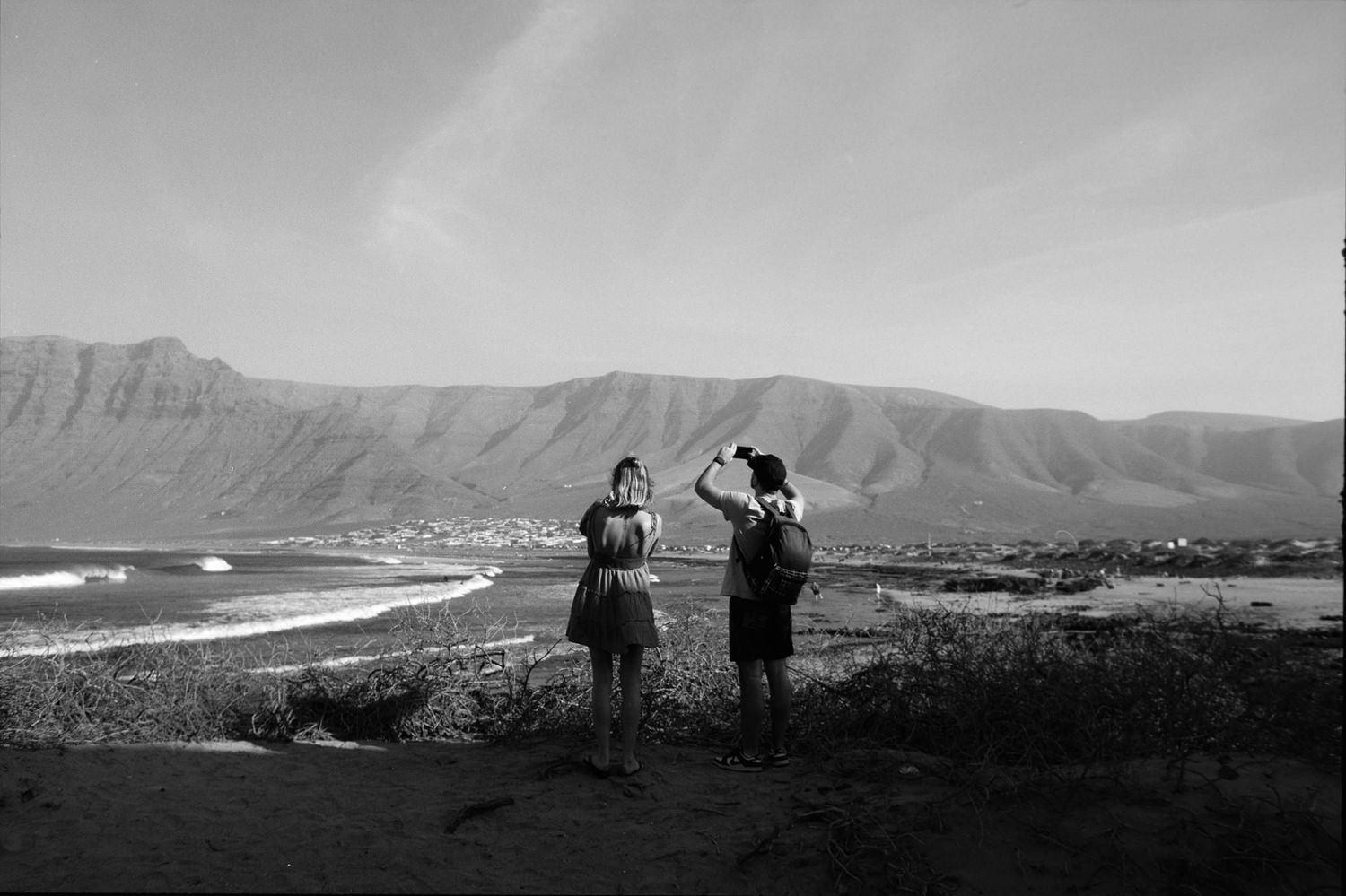 Two figures stand on a hilltop overlooking a scenic coastal landscape with mountains in black and white.