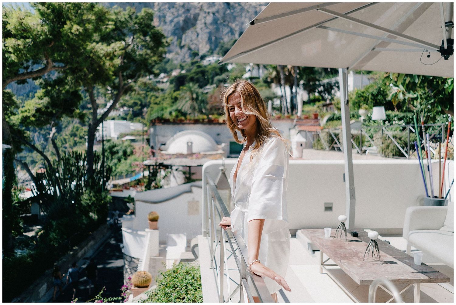 Person in white stands on sunny Mediterranean terrace with outdoor dining setup overlooking coastal scenery.