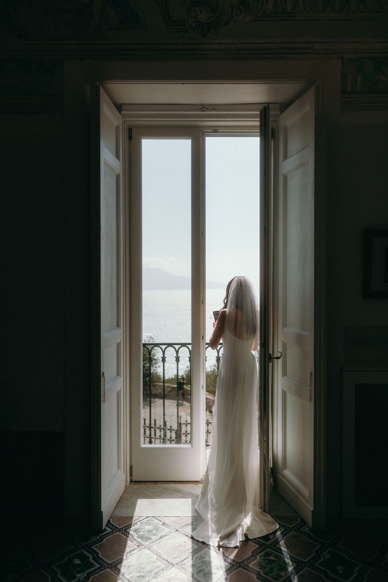 A person in a flowing white dress stands silhouetted in a doorway overlooking a scenic balcony view of mountains and water.