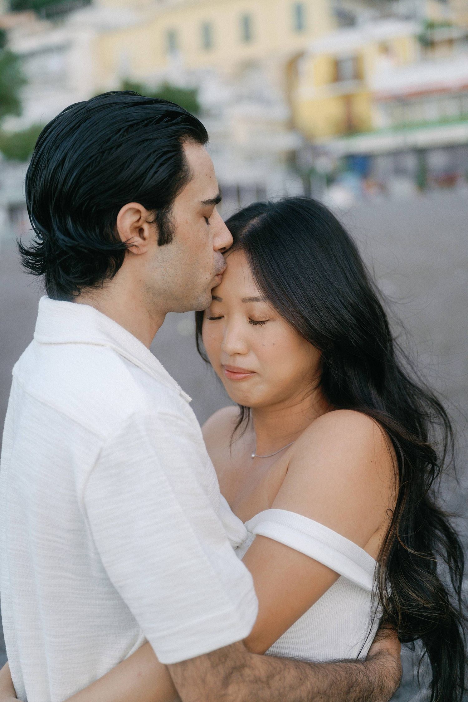 A romantic couple embraces in white outfits against a blurred coastal town background.