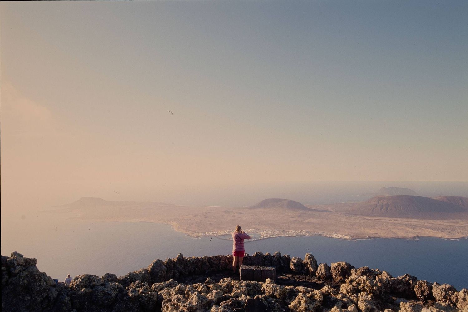 A silhouette stands atop rocky cliffs overlooking distant mountain ranges at dusk.