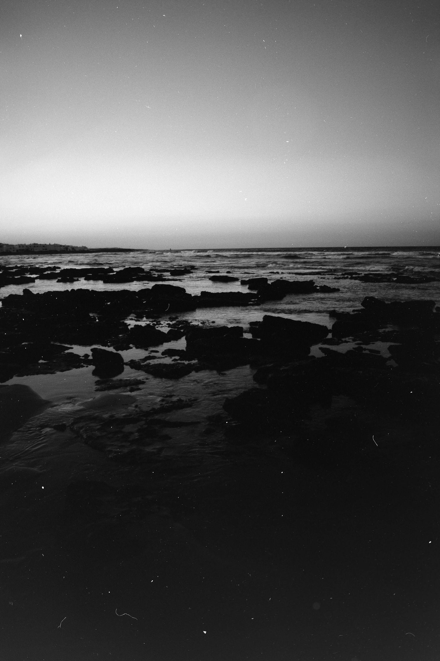 A black and white coastal seascape with rocky tide pools stretching towards a misty horizon at dusk.
