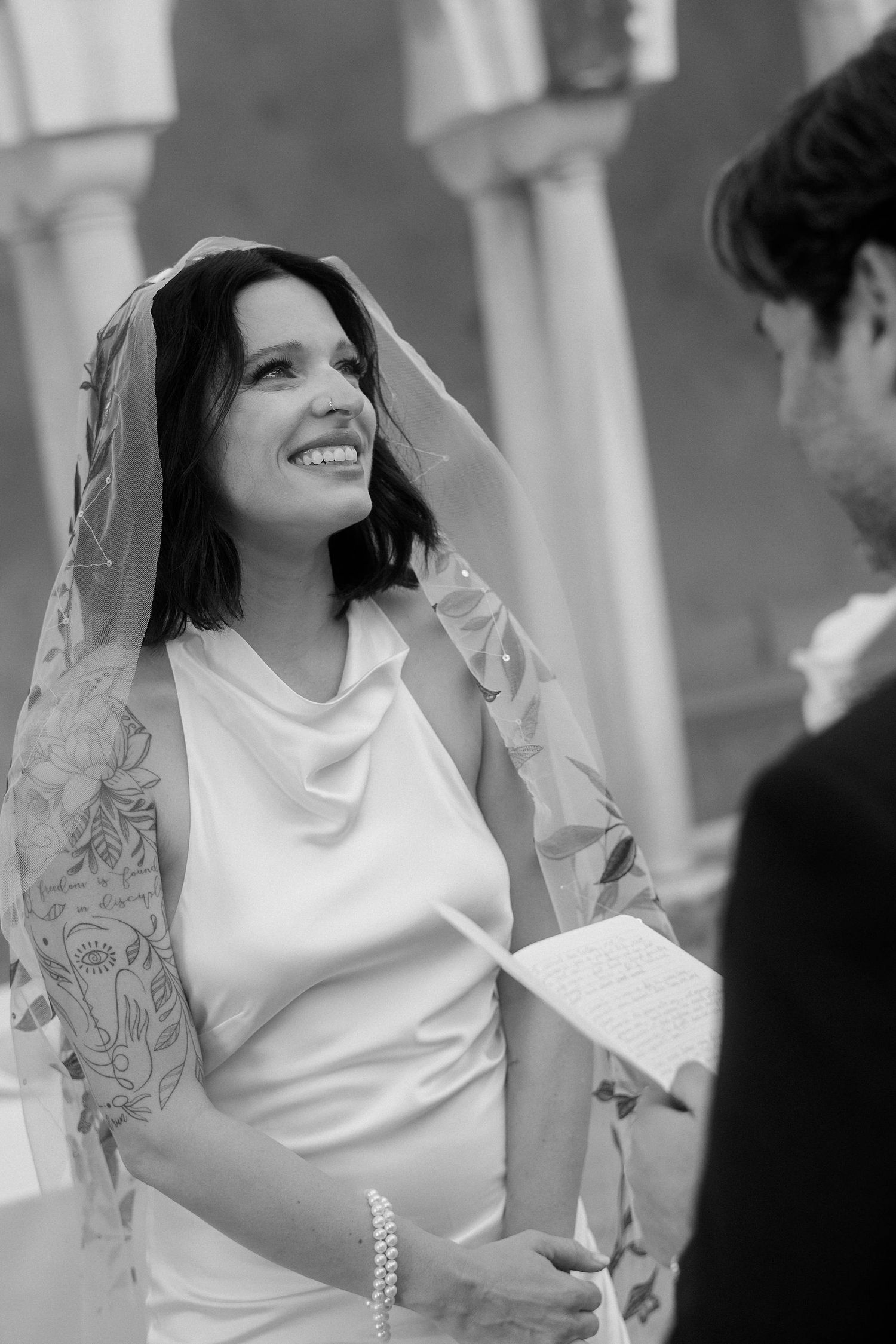 A bride in a white dress and veil smiles joyfully during her wedding ceremony.