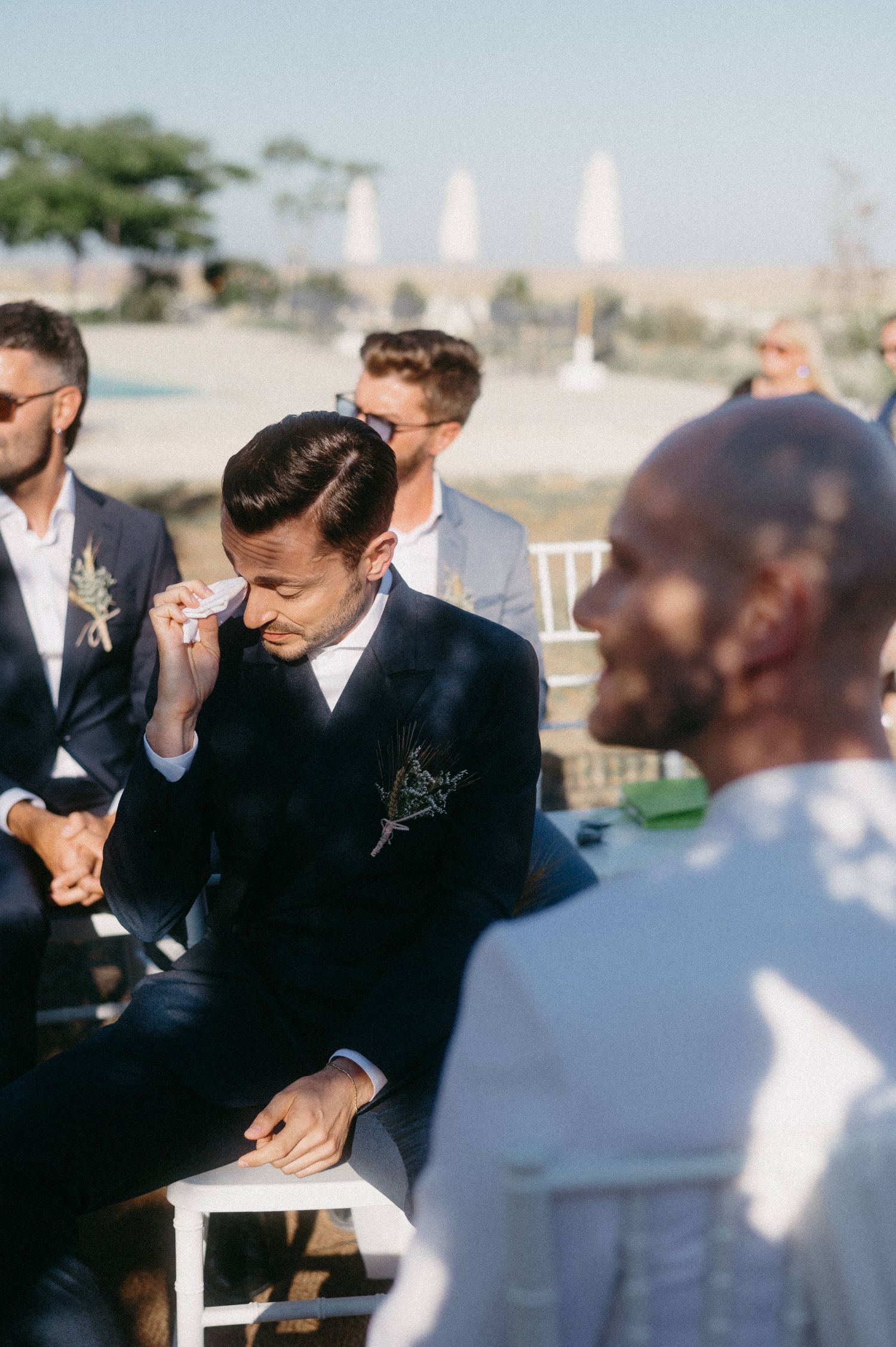 Emotional moment at an outdoor wedding ceremony with guests seated in navy suits and white attire.