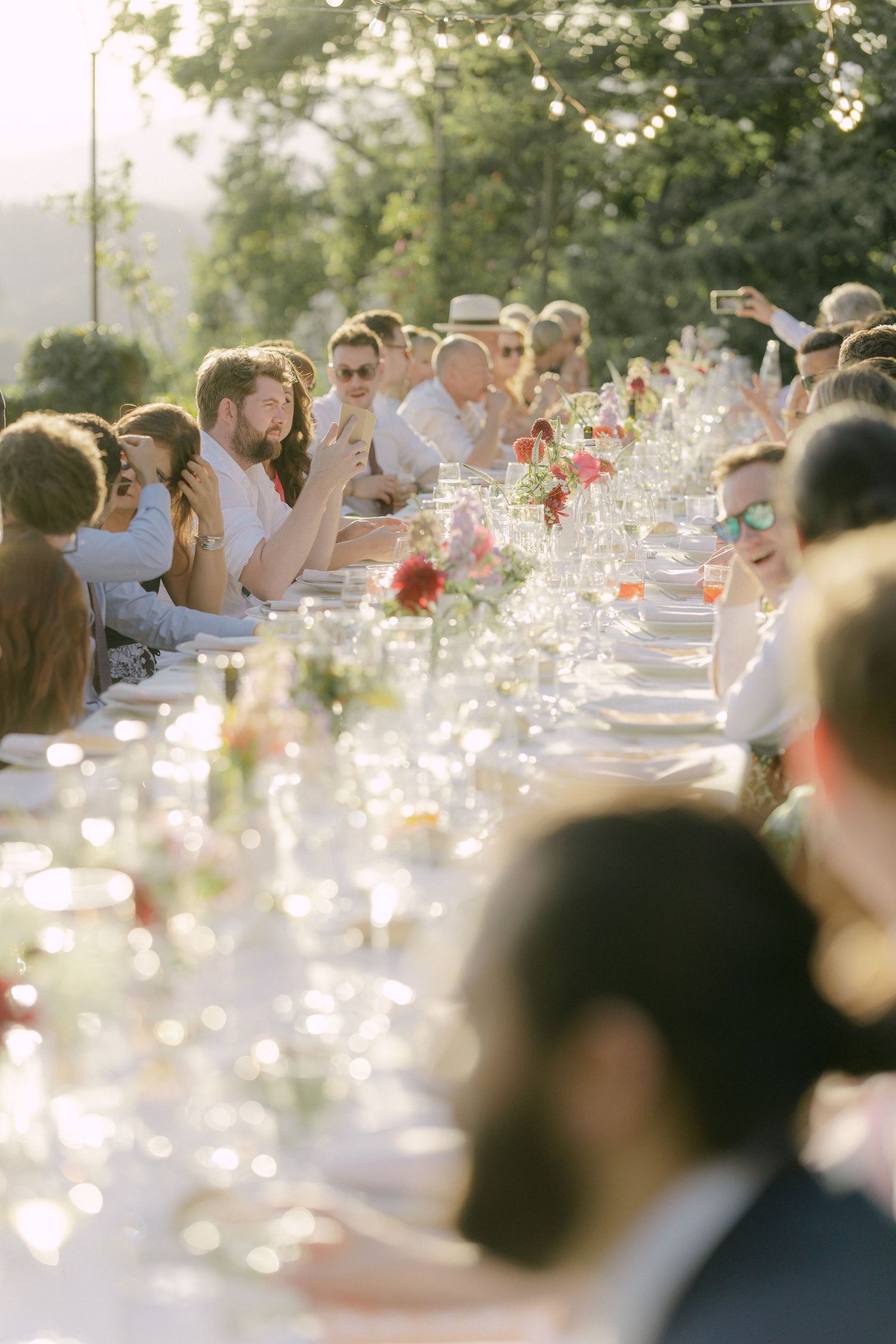 Guests gather at an elegant outdoor dinner party with sunlit tables and glassware creating a magical golden-hour atmosphere.