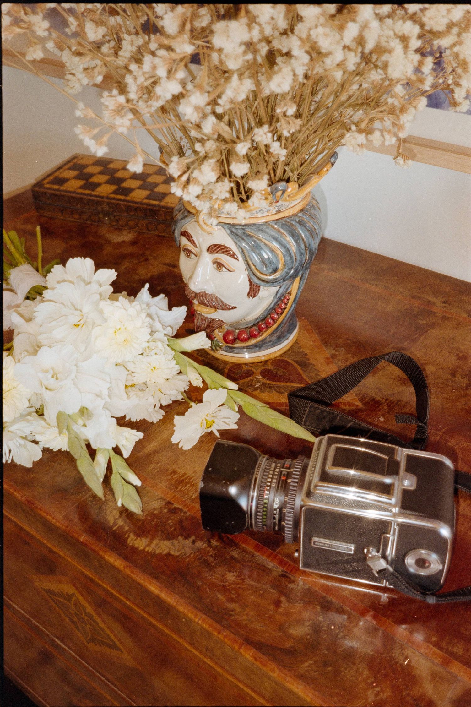 Vintage Hasselblad camera sits on wooden table beside decorative vase with dried flowers and white blooms.