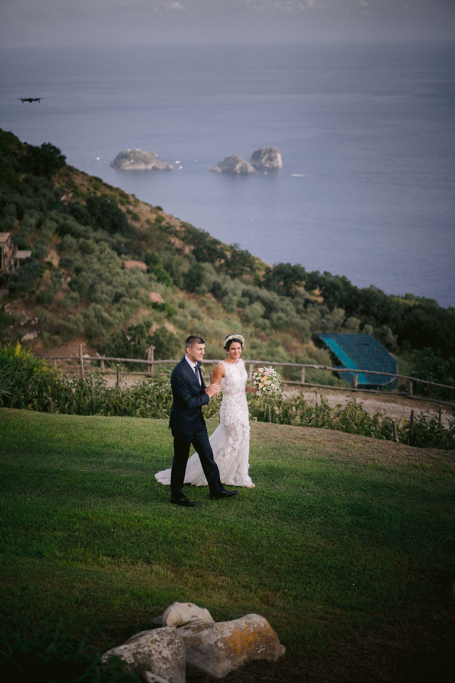 A couple walks on a grassy hillside overlooking rocky ocean cliffs and coastline at sunset.