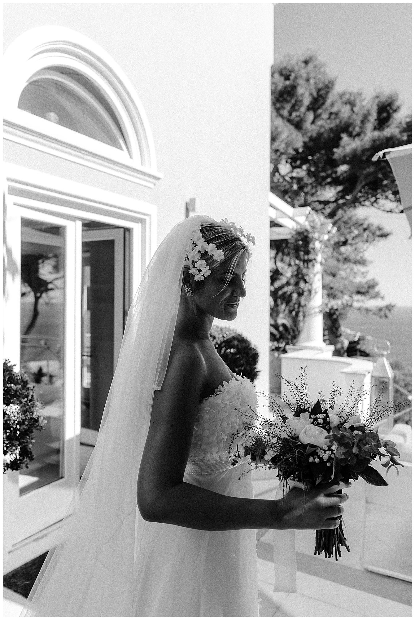 Black and white profile of person in wedding dress and veil holding bouquet by arched doorway.