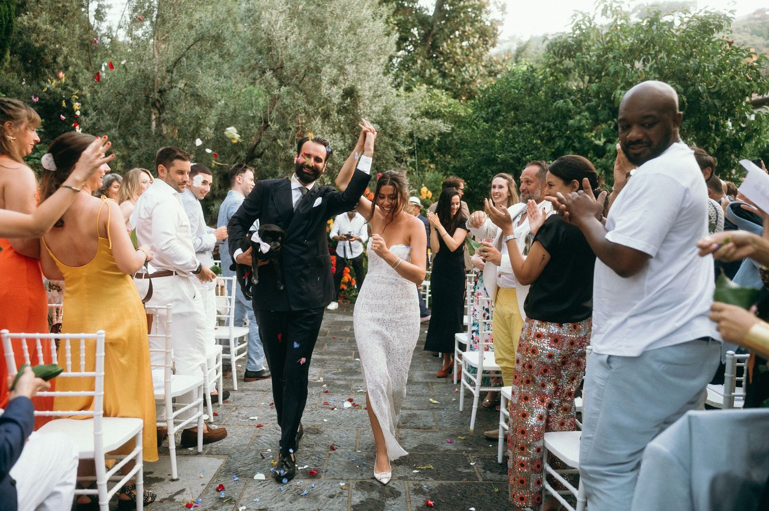 A lively celebration with guests cheering as the newlyweds joyfully walk down an outdoor aisle lined with white chairs.