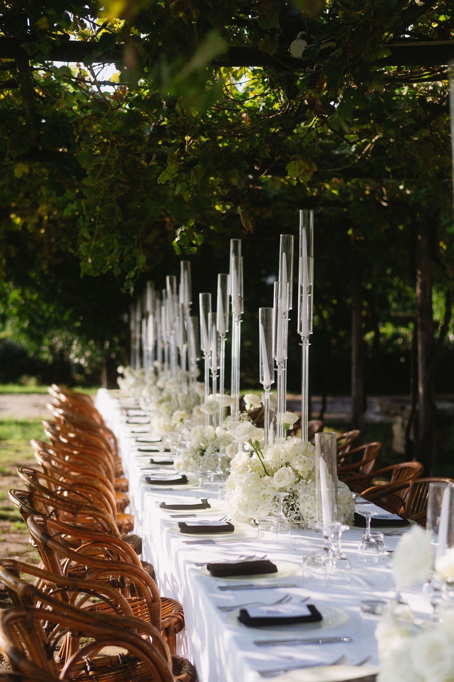 Elegant outdoor wedding reception table setting with white linens, tall candles, and natural wooden chairs under lush greenery.