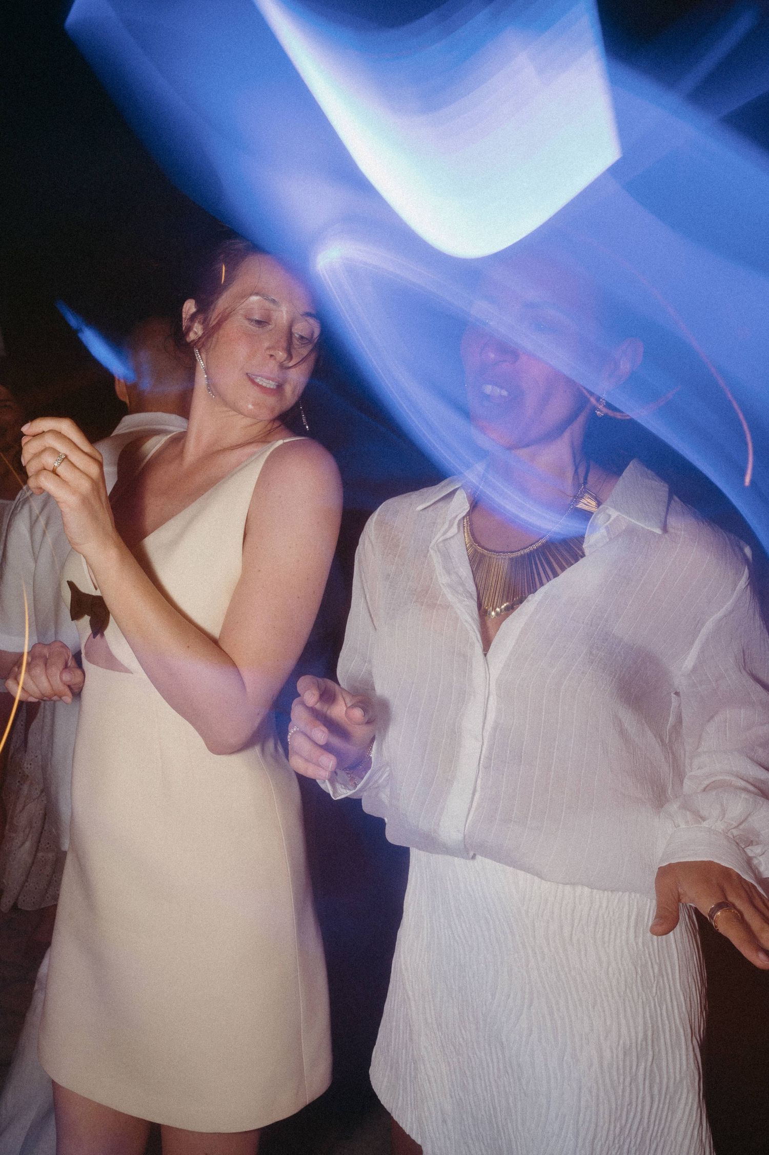 A couple dancing together at a party under blue lighting while wearing white and cream colored outfits.
