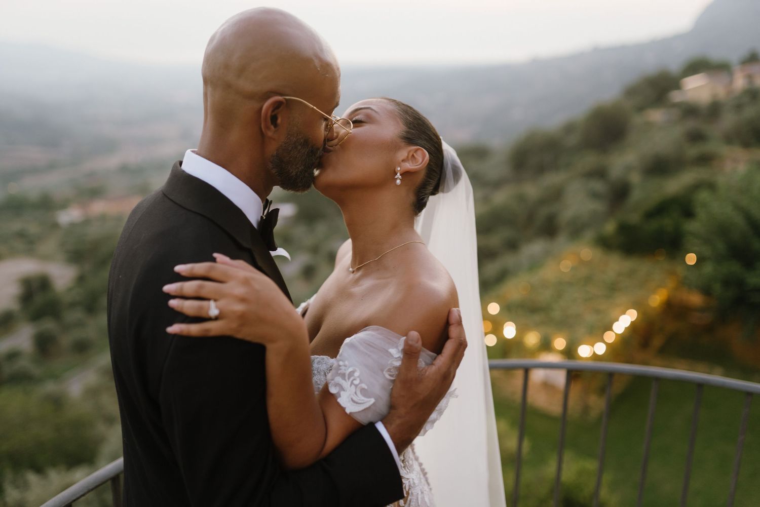 A romantic wedding couple shares a tender kiss against a scenic mountain backdrop at sunset.