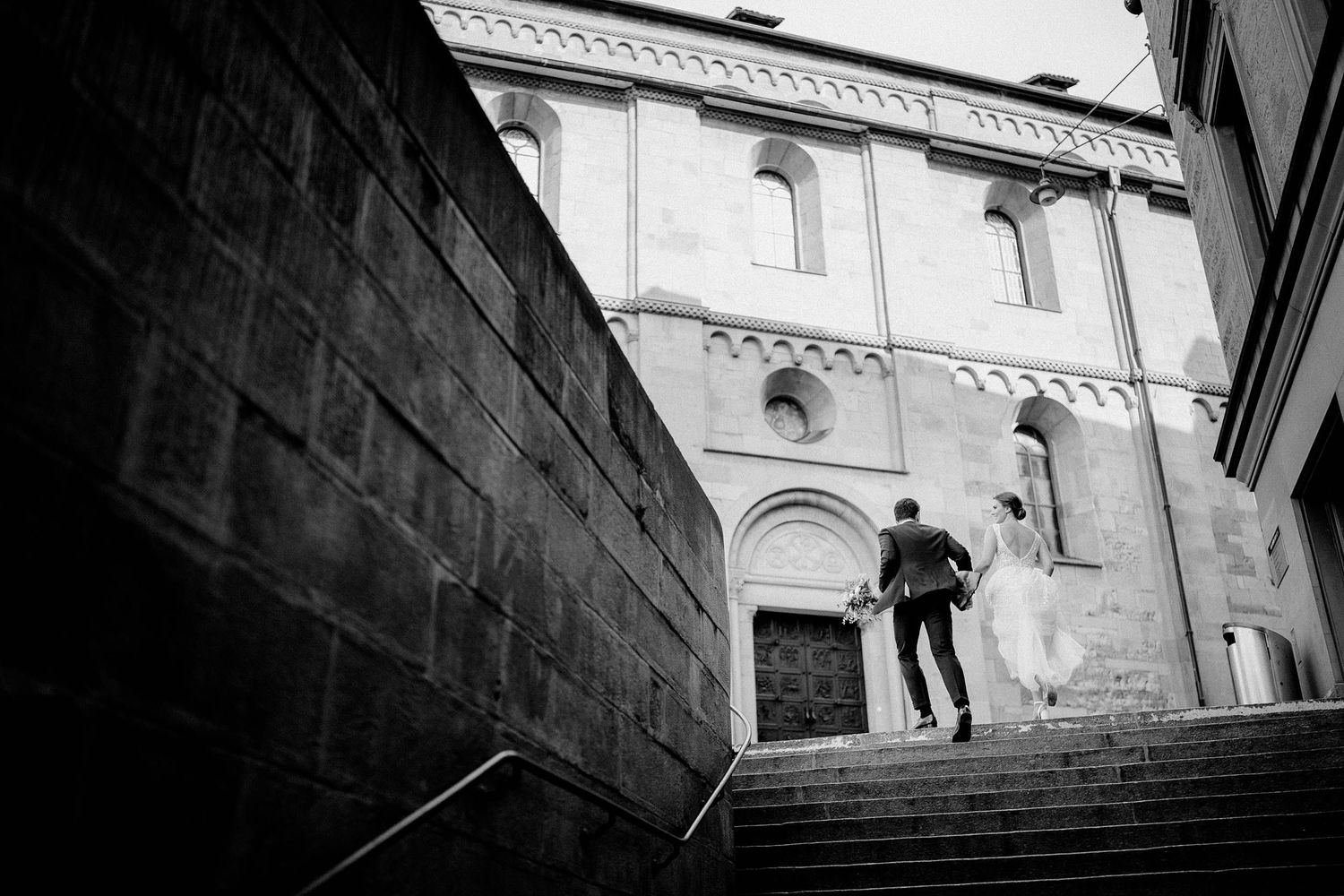 Black and white architectural photograph showing a couple ascending stone steps beside a tall church wall.