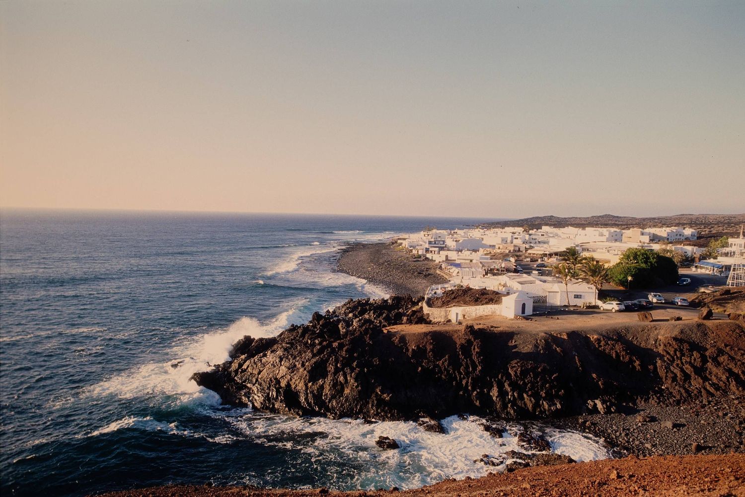 Coastal landscape with white buildings perched on rugged volcanic cliffs overlooking the ocean at sunset.