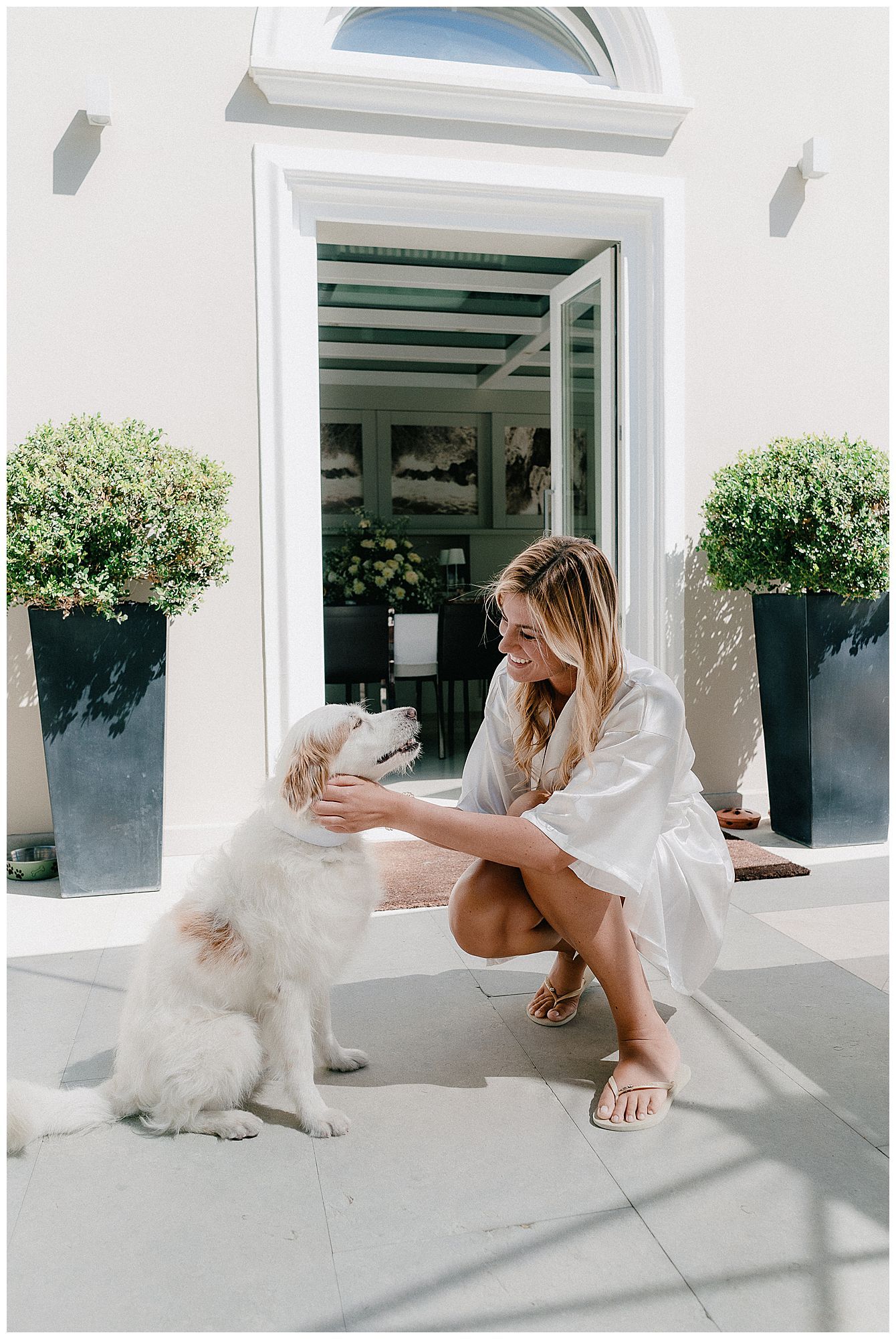 A person in a white outfit kneels on a porch to pet a fluffy white dog beside two large planters at an elegant entrance.
