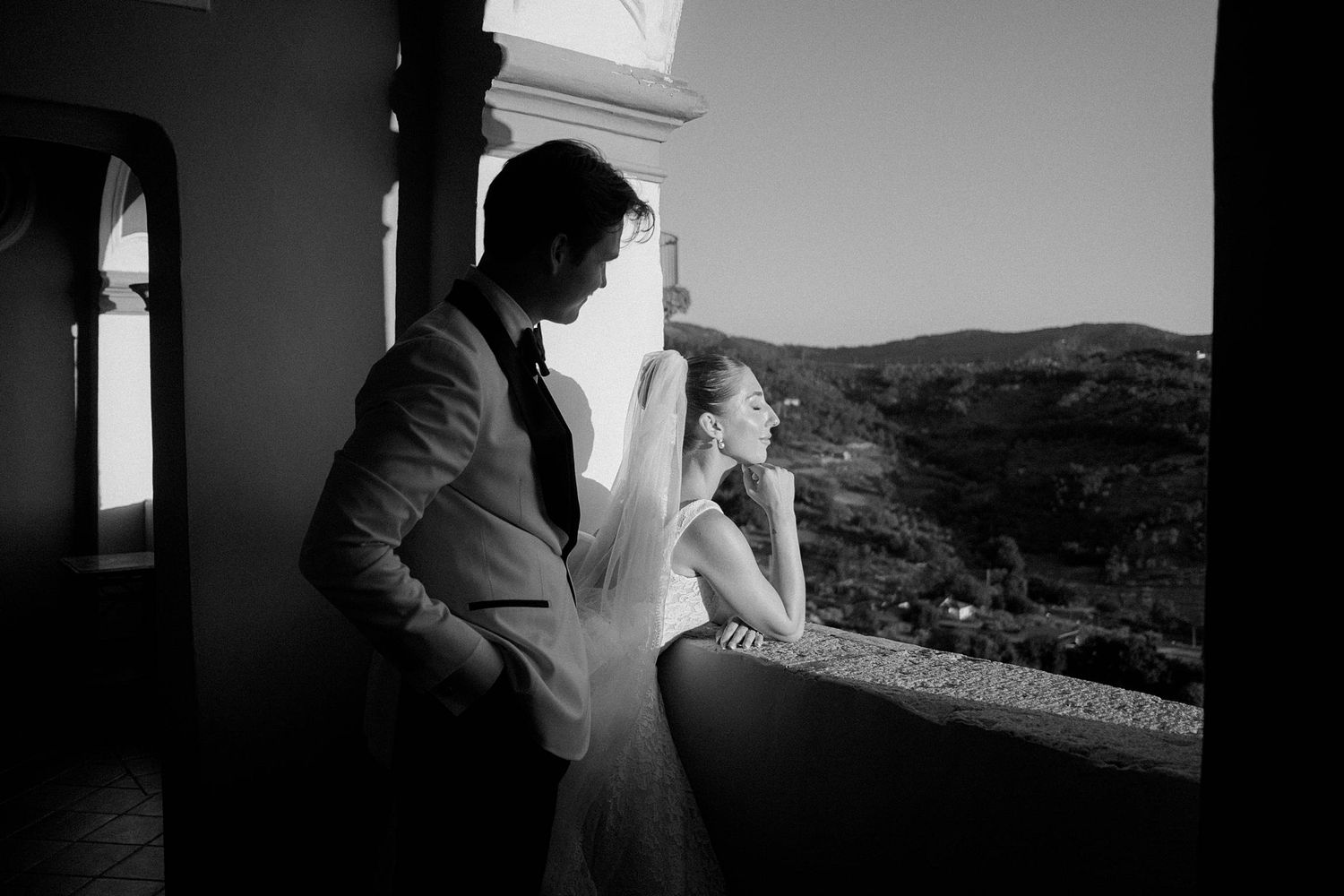 Black and white photo of a bride and groom gazing at scenic hills from a sunlit balcony on their wedding day.