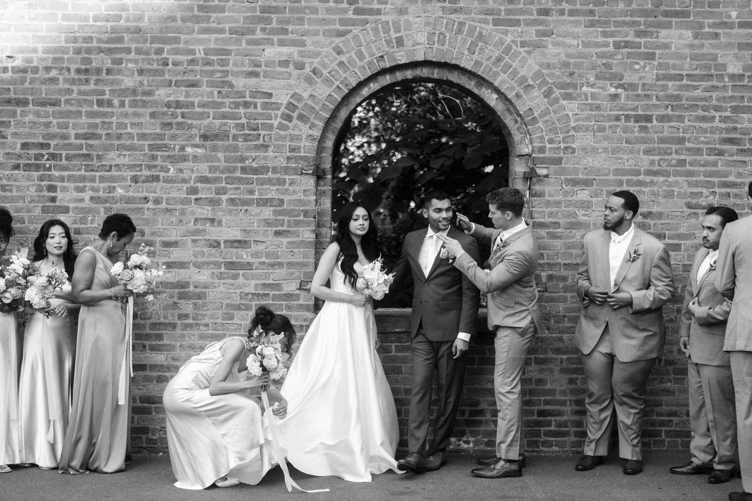 A panoramic wedding party lineup poses against a rustic brick wall in black and white formal attire.