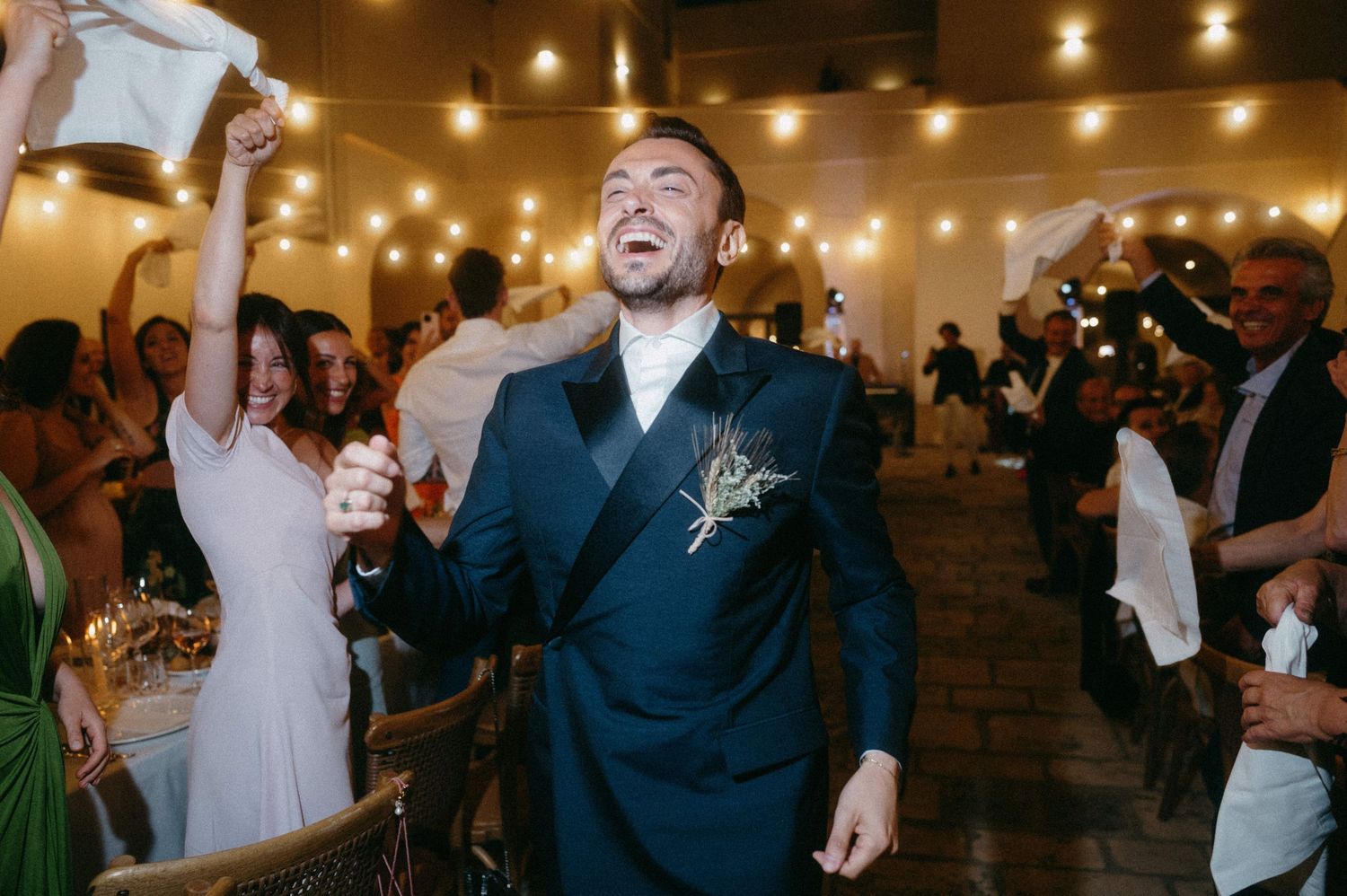Wedding guests dancing and celebrating on a dimly lit dance floor under string lights at an evening reception.