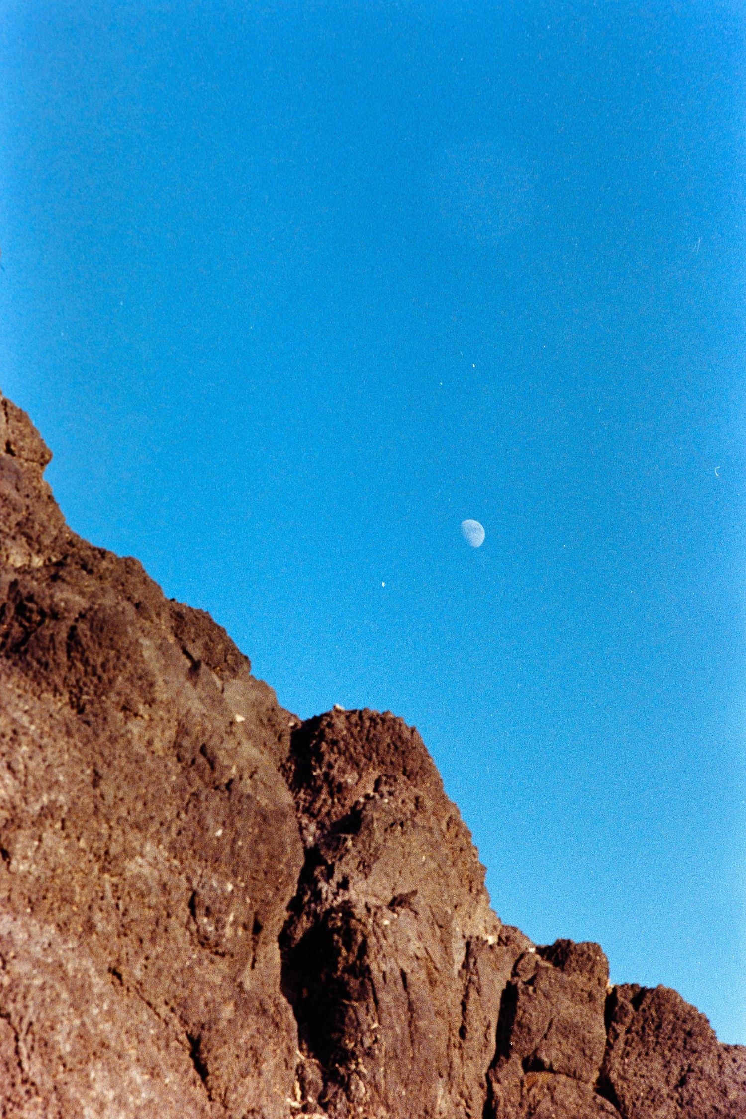 Moon visible in bright blue sky over rugged brown mountain peaks in desert landscape against clear horizon.