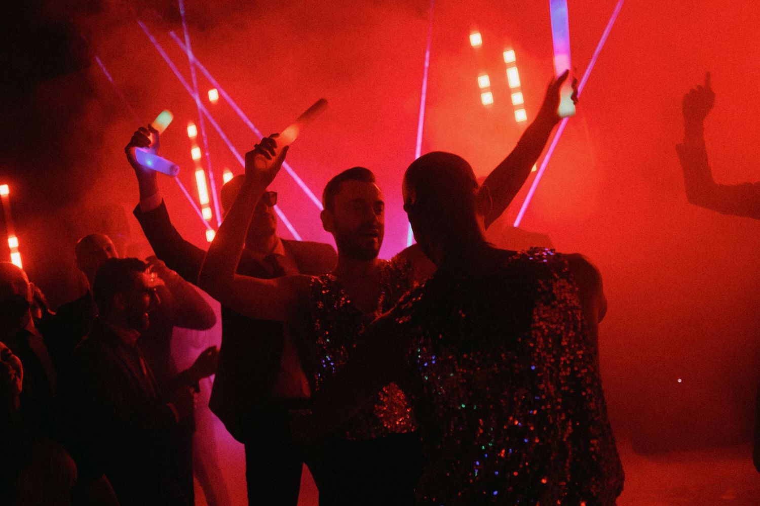 Silhouetted crowd dancing with raised arms in a nightclub illuminated by bright red lighting and fog effects.