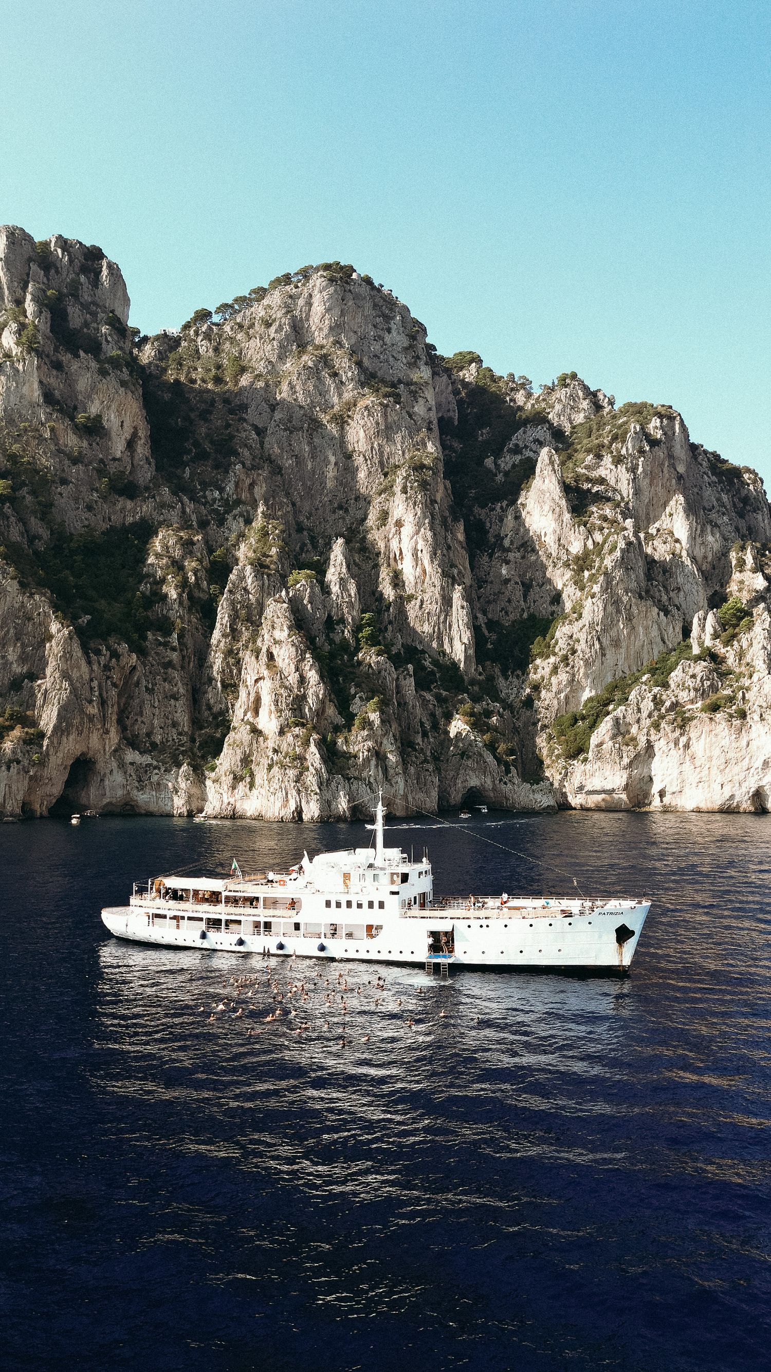 A white luxury yacht cruises past dramatic limestone cliffs along the Mediterranean coastline.