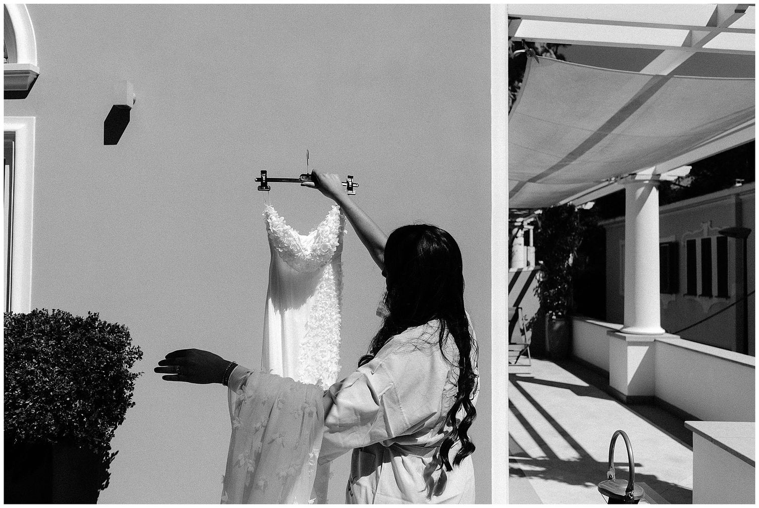 Someone in long sleeves hangs up a white wedding dress against a light colored wall outdoors.