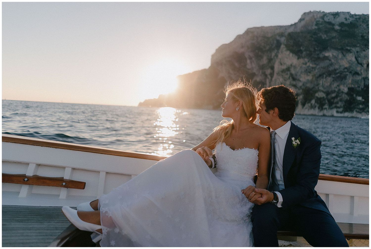 Newlywed couple embracing on a yacht with coastal cliffs and sunset over Mediterranean waters in background.
