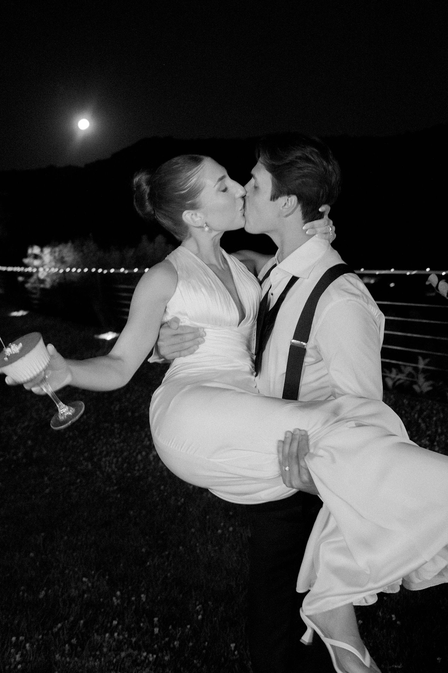 Black and white photo of groom carrying bride while kissing under moonlit night sky at outdoor wedding reception.
