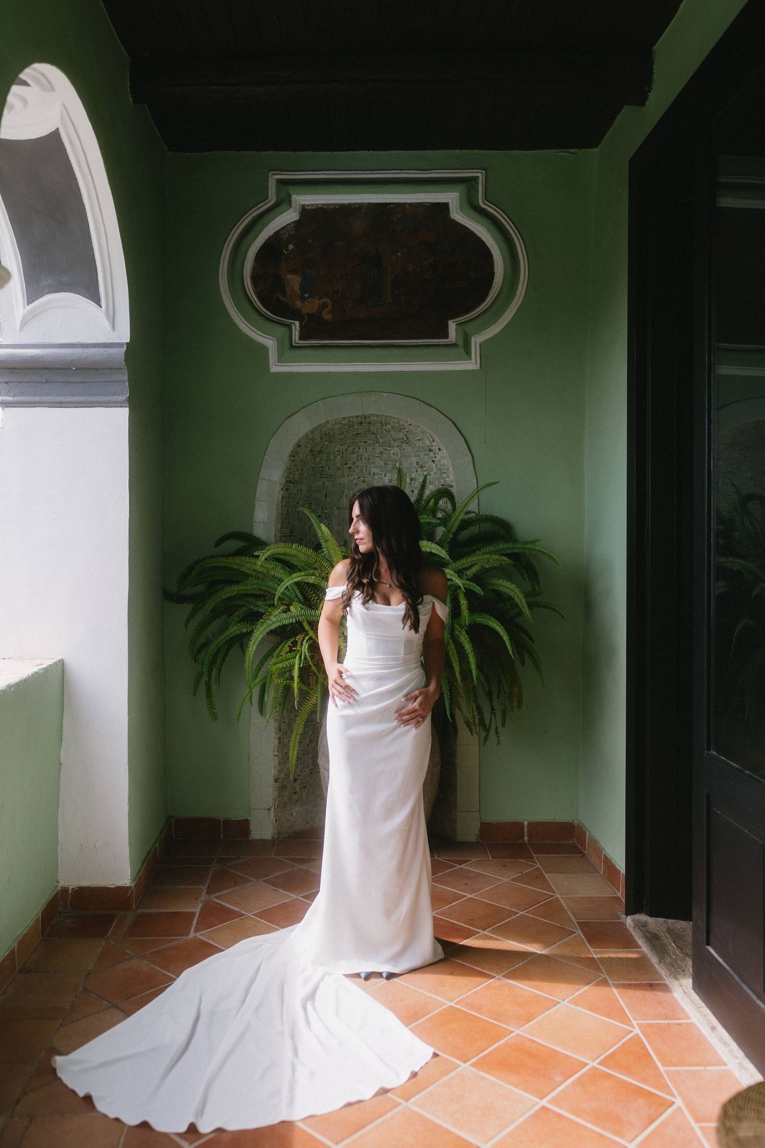 A person in a flowing white dress sits in a sunlit corridor with green walls and terracotta flooring.